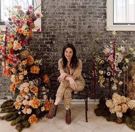 A woman sitting on a bench surrounded by large floral arrangements in an indoor setting with a brick wall background.