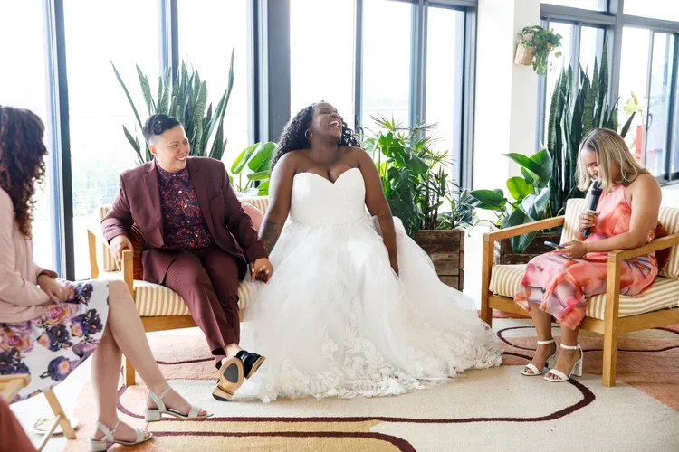 A queer couple including a bride in a white wedding dress sitting with guests, laughing and smiling in a bright room with large windows and green plants. One woman is holding a microphone.