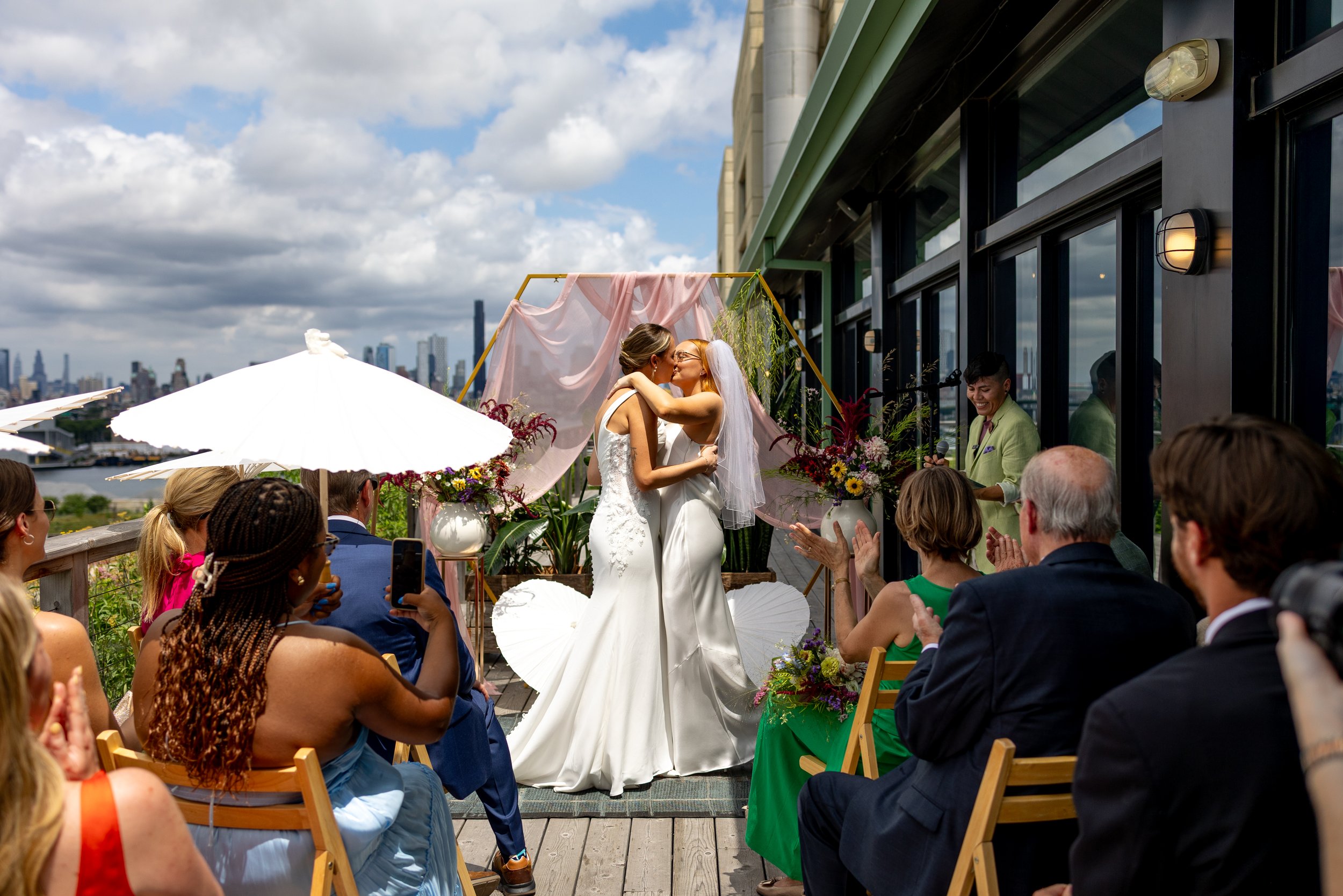 Queer wedding ceremony on a rooftop with two brides kissing, surrounded by seated guests, city skyline in the background, decorated with flowers and drapes.
