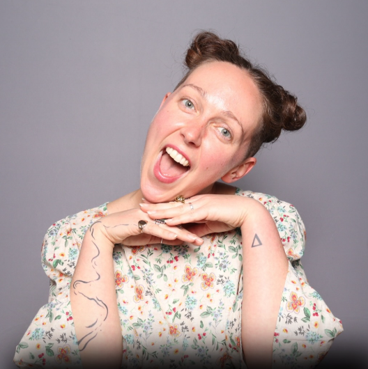Joyful Brooklyn wedding photographer wearing floral top and braided buns, smiling with hands folded under chin.