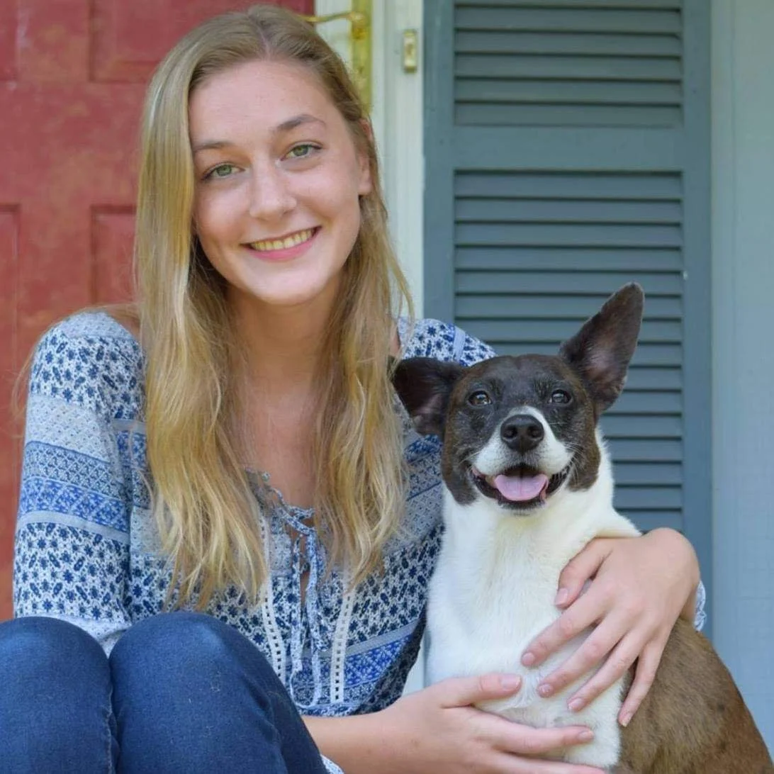 A young woman with long blonde hair smiling and sitting on the ground, holding a small black and white dog with one ear up and one down, outside in front of a red door and blue shutters.