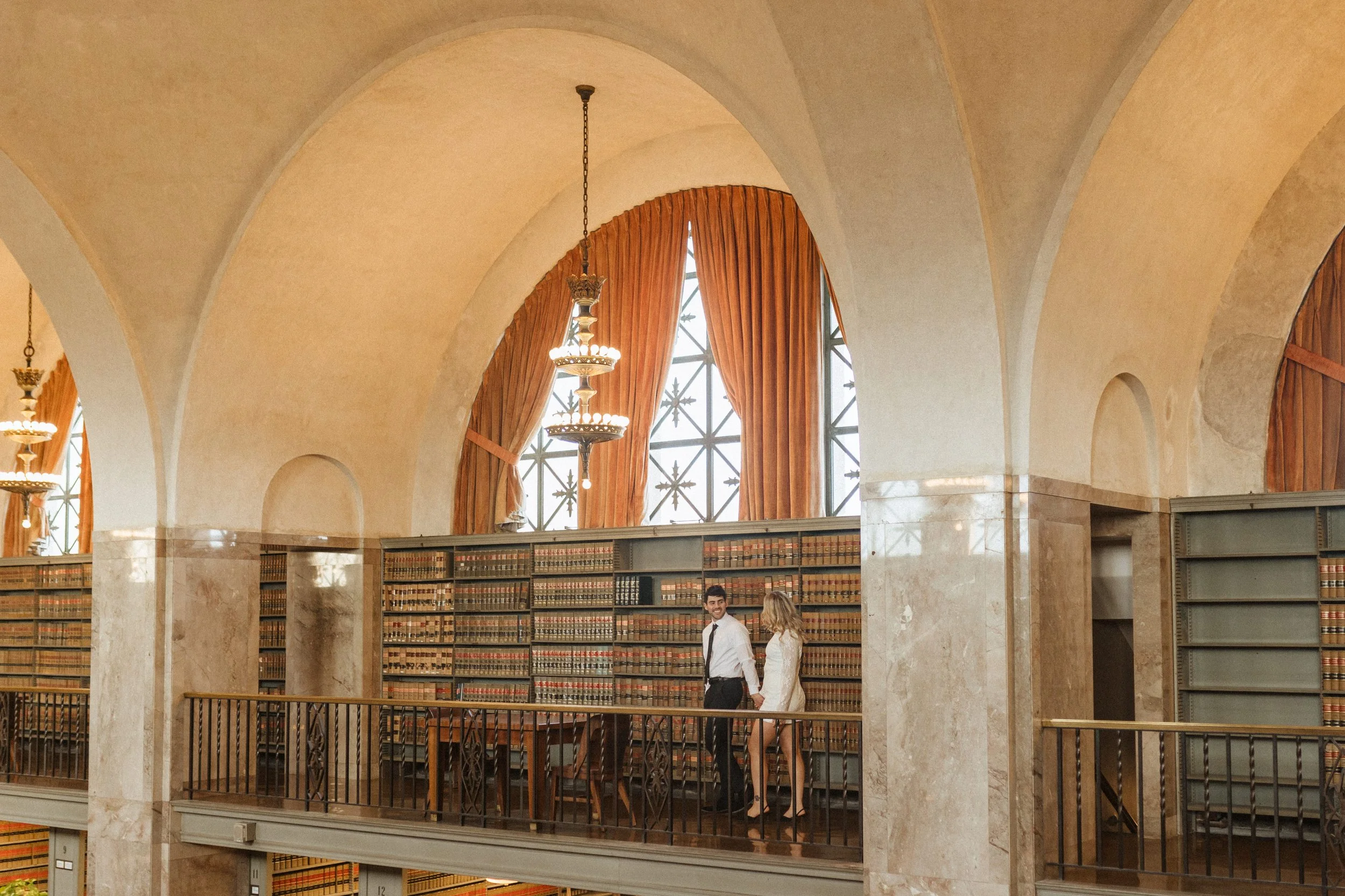 Inside a grand, historic library with high arched ceilings, large windows with orange curtains, and chandeliers. Two people are standing near bookshelves, engaged in conversation.