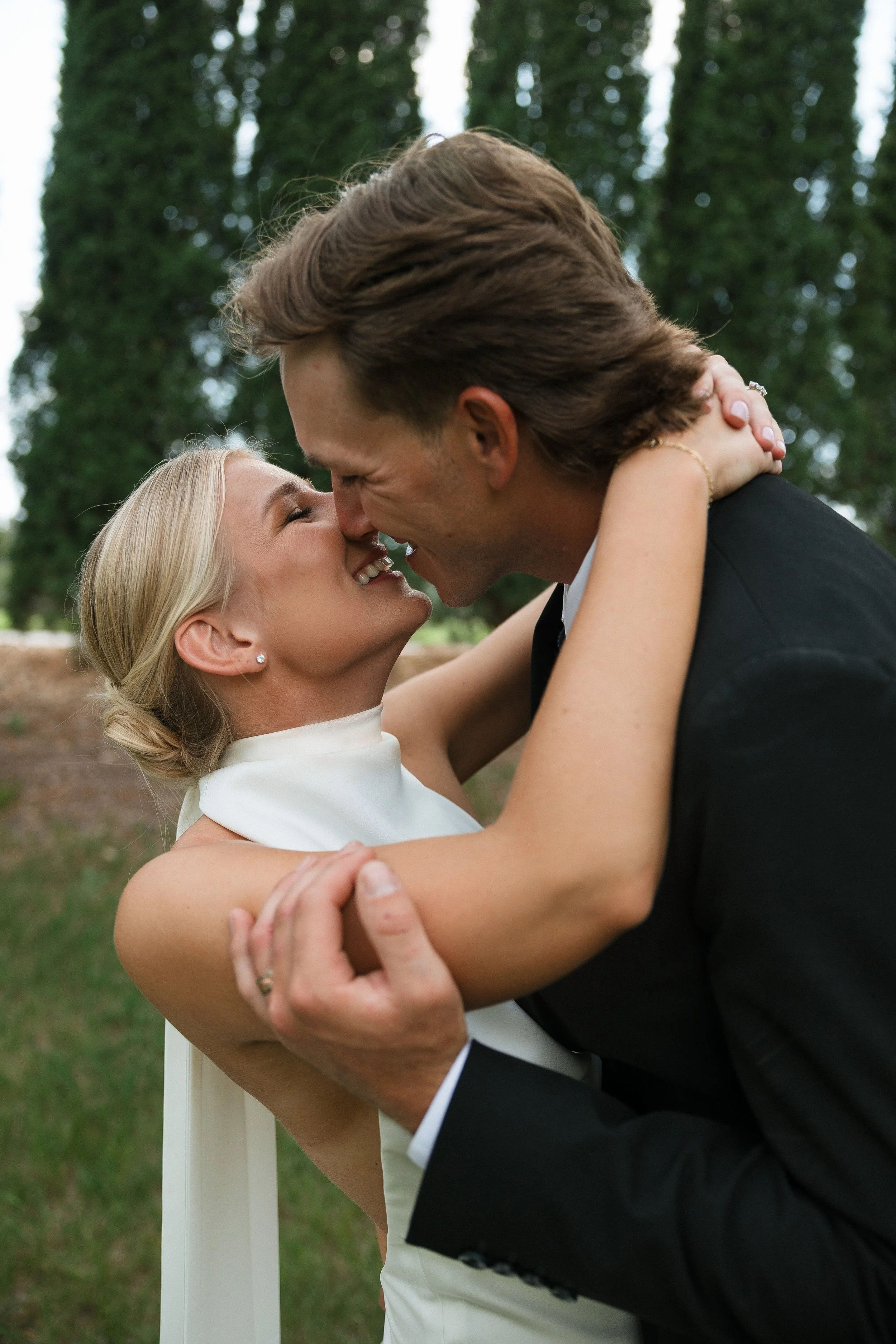 A couple is embracing outdoors, about to kiss, with trees in the background.
