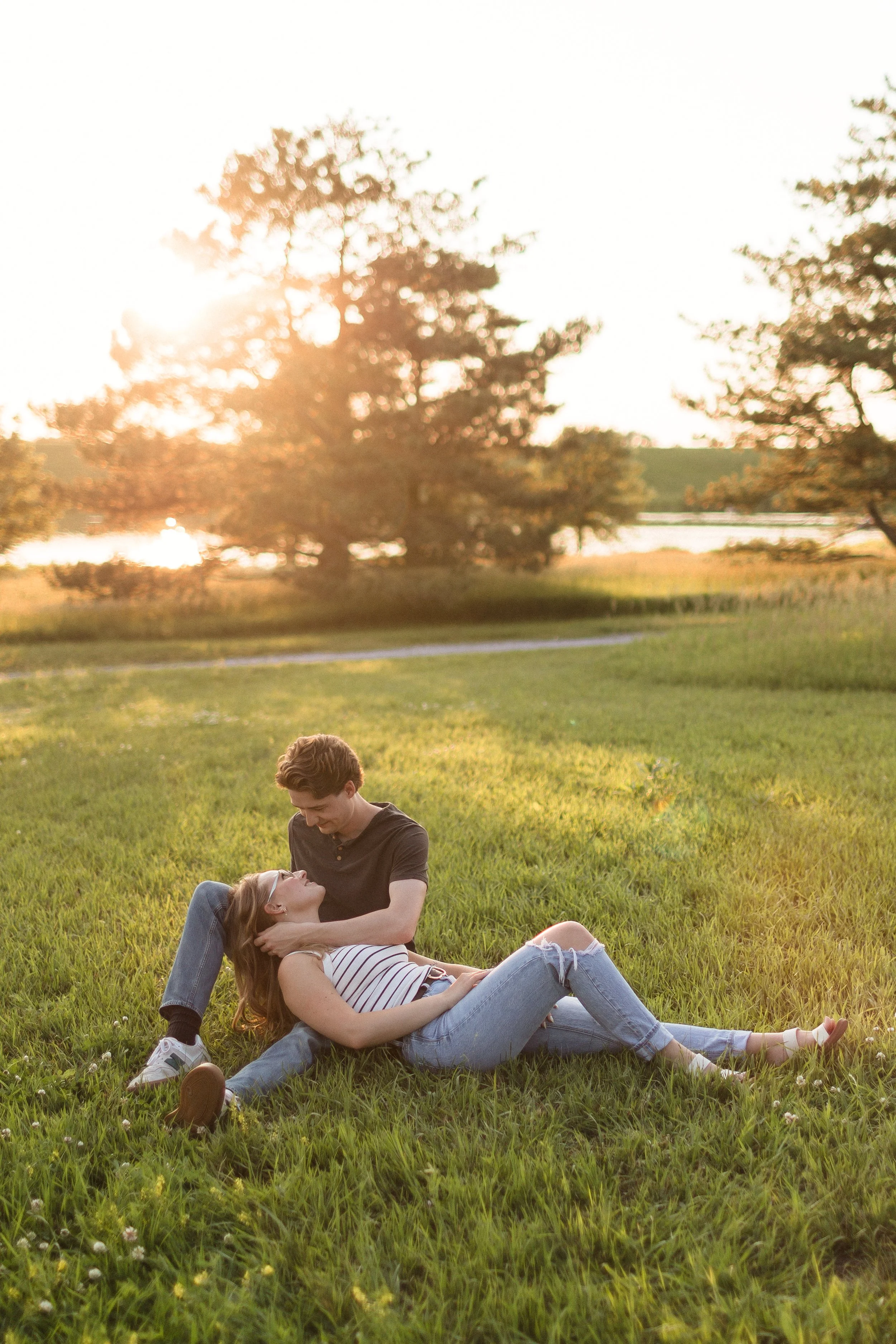 A young couple sitting on grass in a park during sunset, with trees and a body of water in the background.