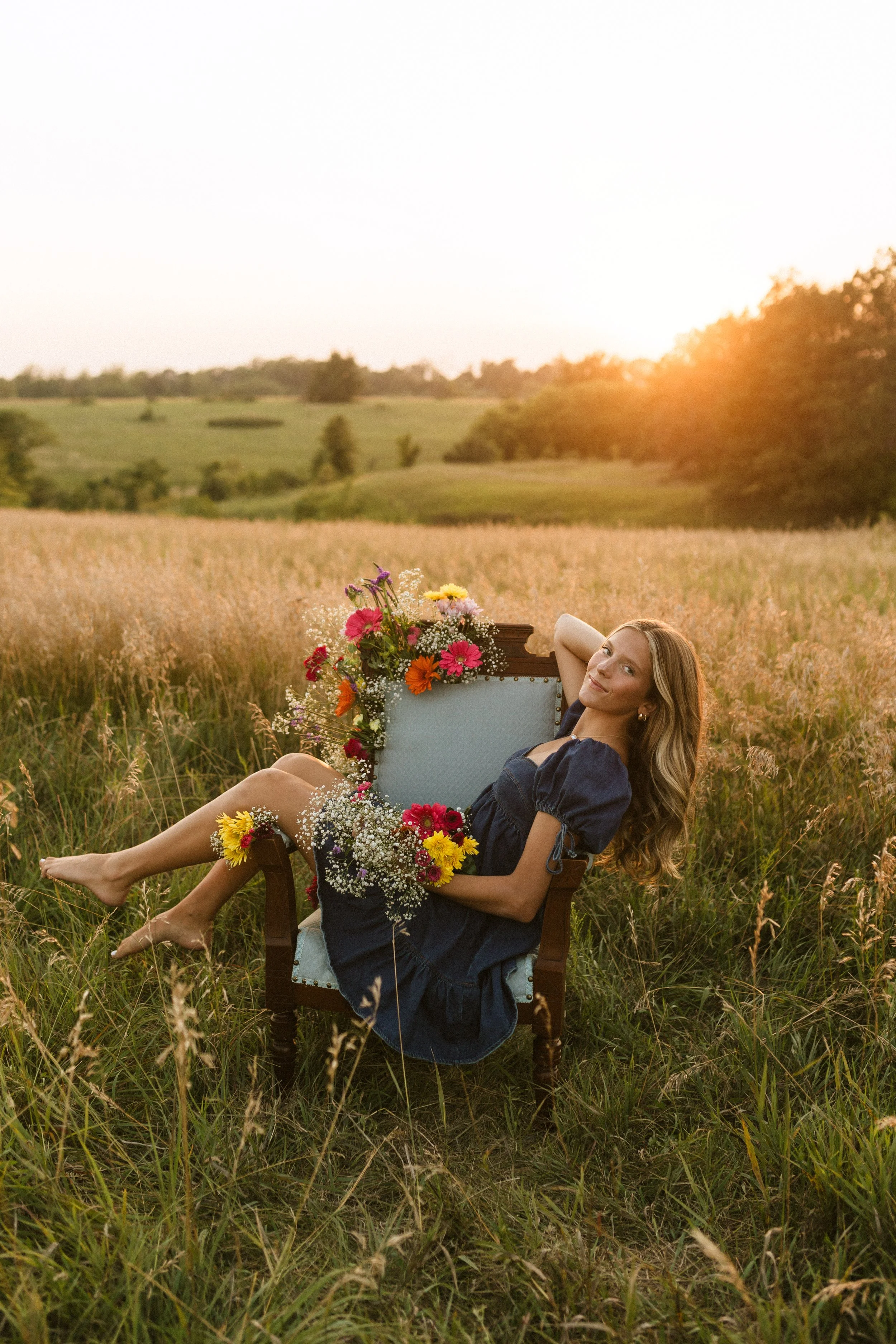 A woman in a navy dress reclining on a vintage chair in a field of tall grass with flowers, with a sunset in the background.