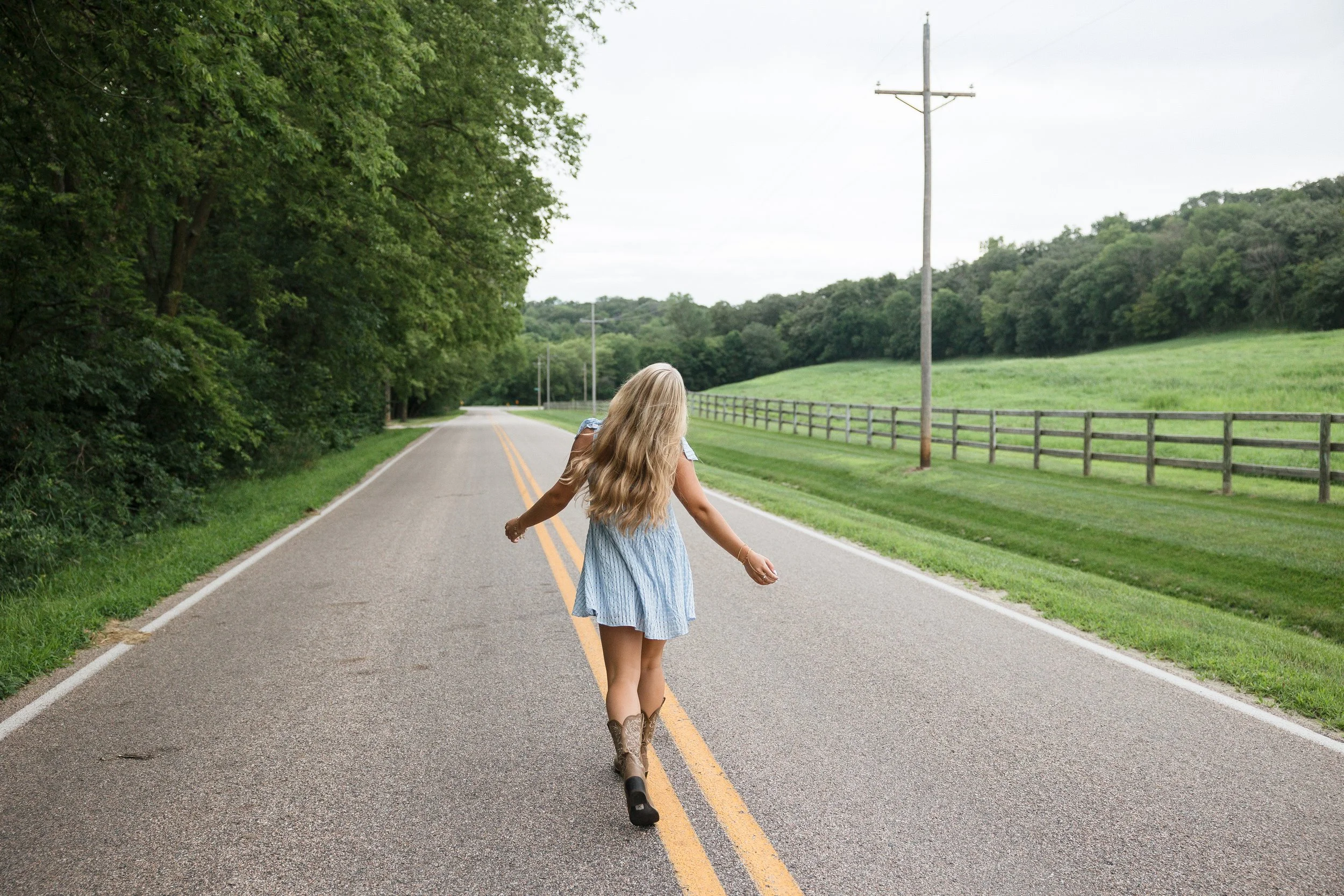 A girl with long blonde hair, wearing a blue dress and cowboy boots, walking on a rural road with double yellow lines, surrounded by green trees and fields under a cloudy sky.