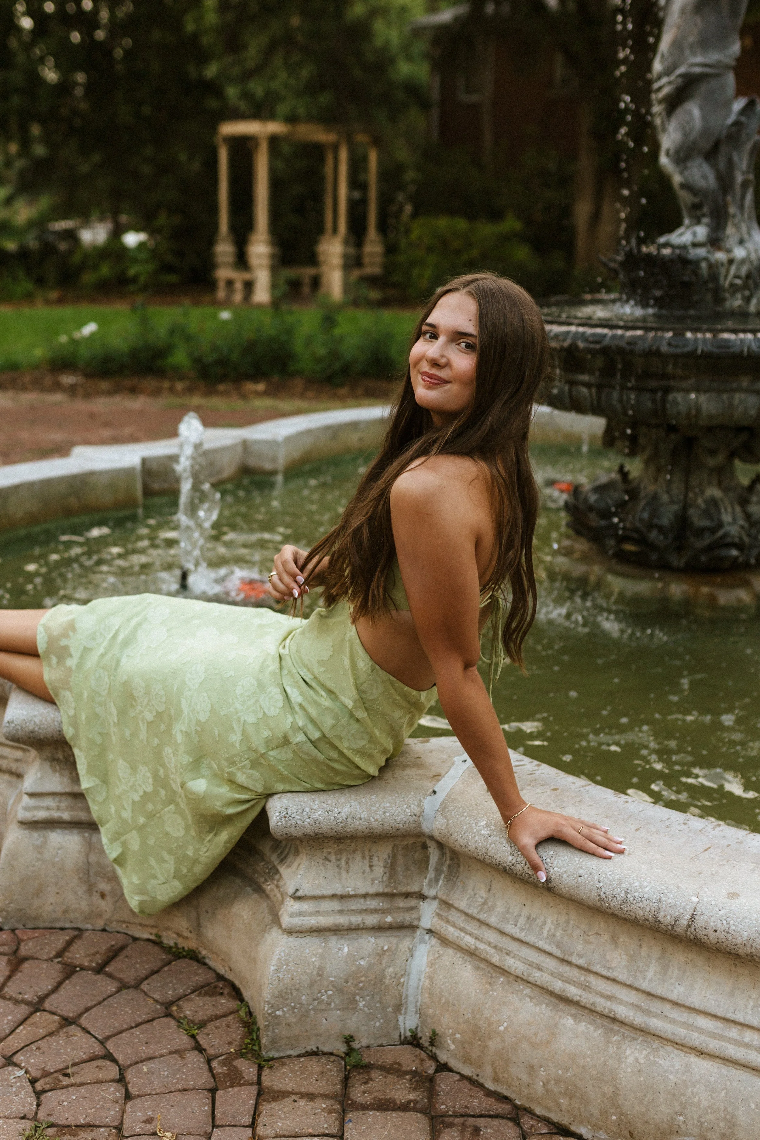 A young woman with long dark hair relaxing on a stone fountain ledge near a pond with a tiered fountain in a lush outdoor garden.