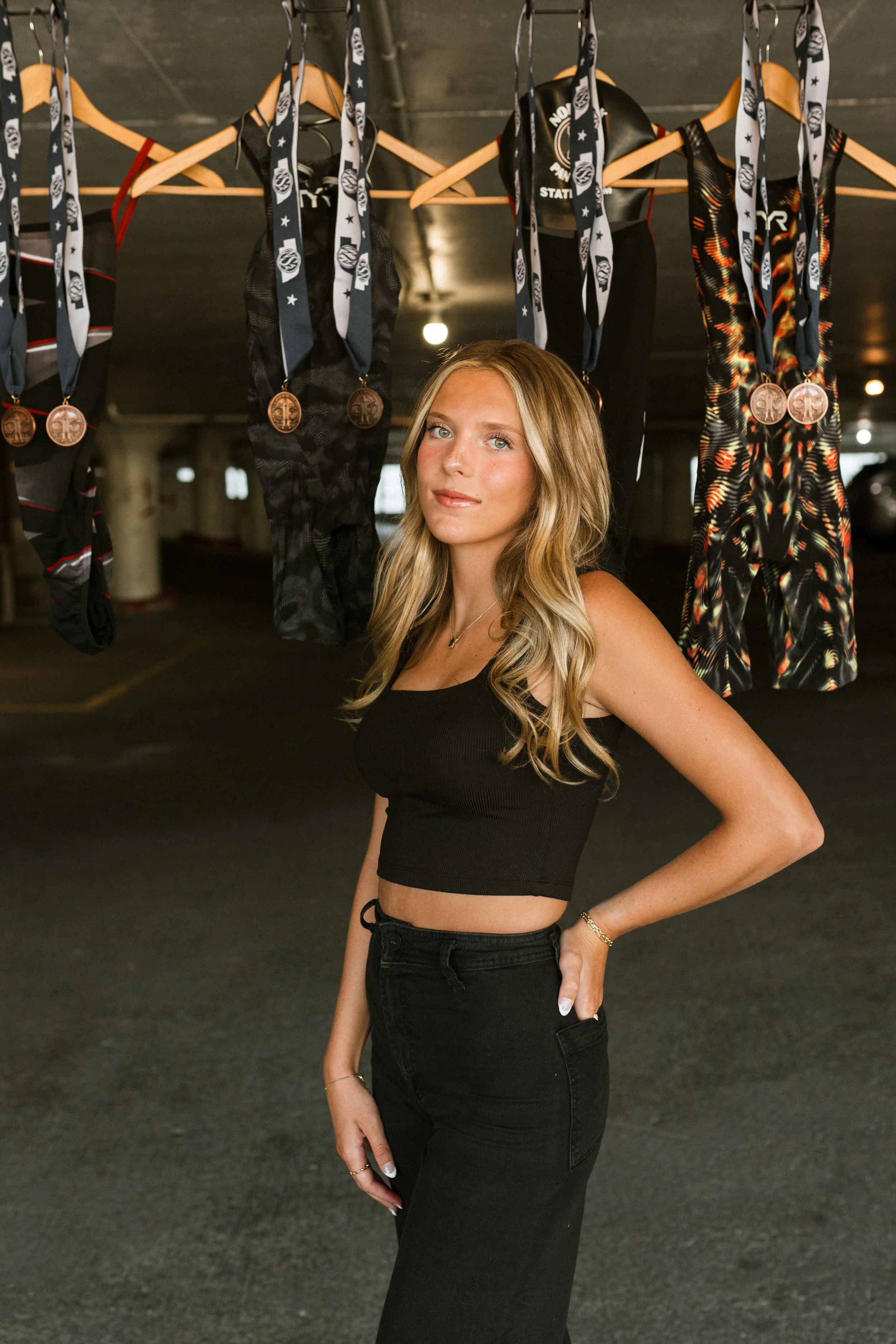 A young woman with long blonde hair in a black crop top and black pants stands in an underground parking garage. Behind her, several medals hang from ribbons on a horizontal bar, with some athletic clothing hanging on hangers. The lighting is dim and industrial.