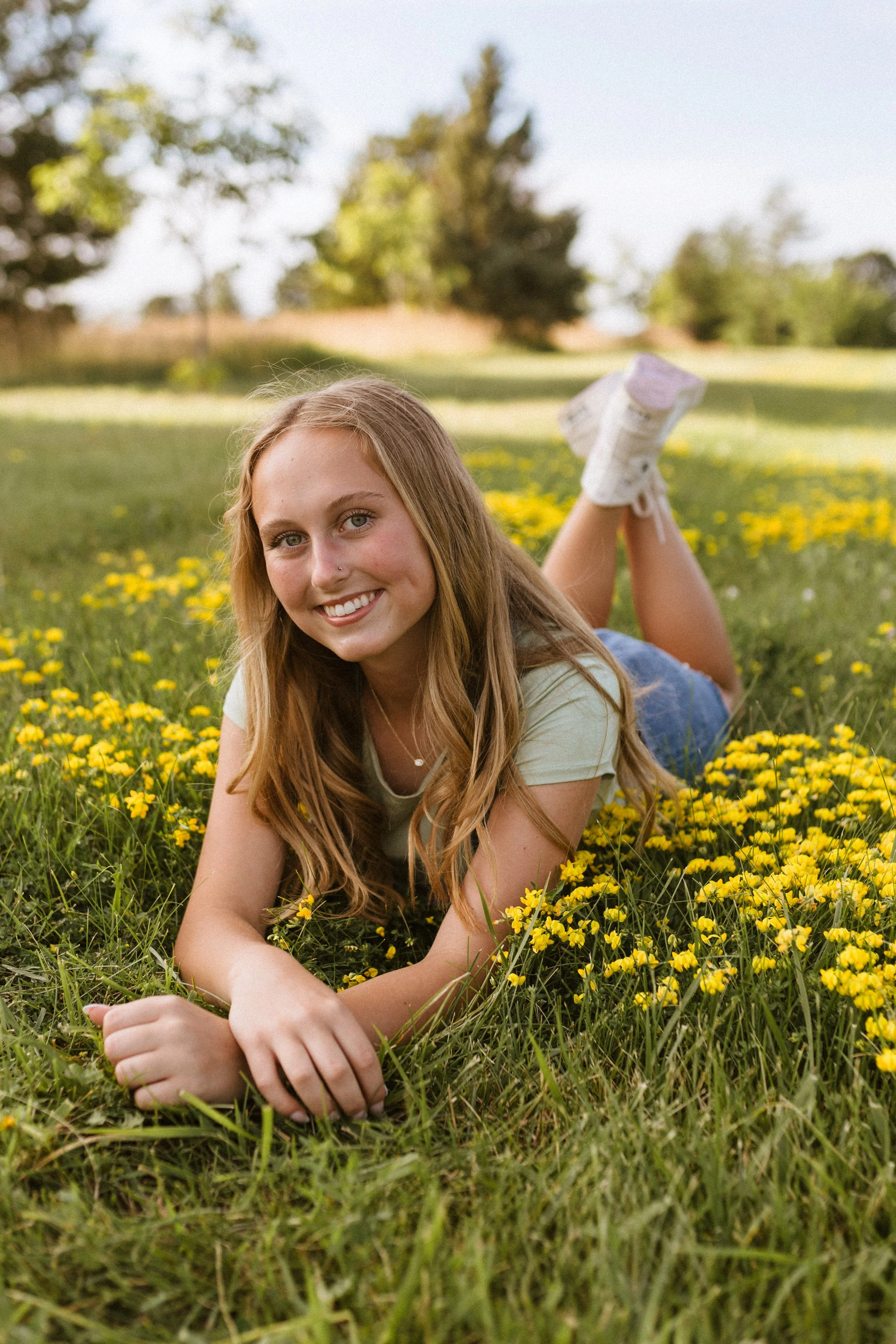A young woman with long, wavy, light brown hair lying on her stomach in a field of yellow flowers, smiling at the camera with a clear sky and trees in the background.