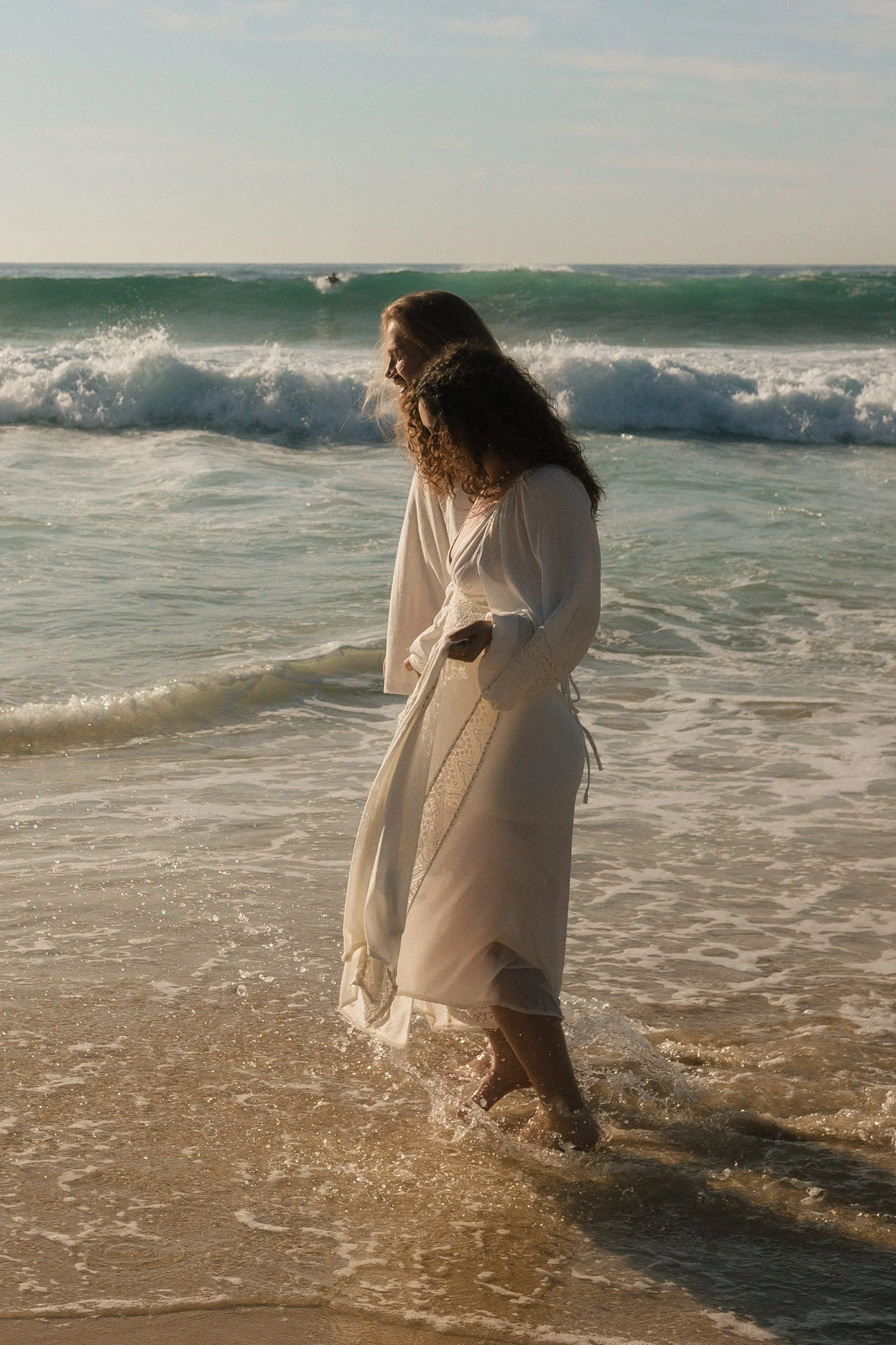 Two women walking in the ocean at sunset, wearing white dresses, with waves crashing around their feet.