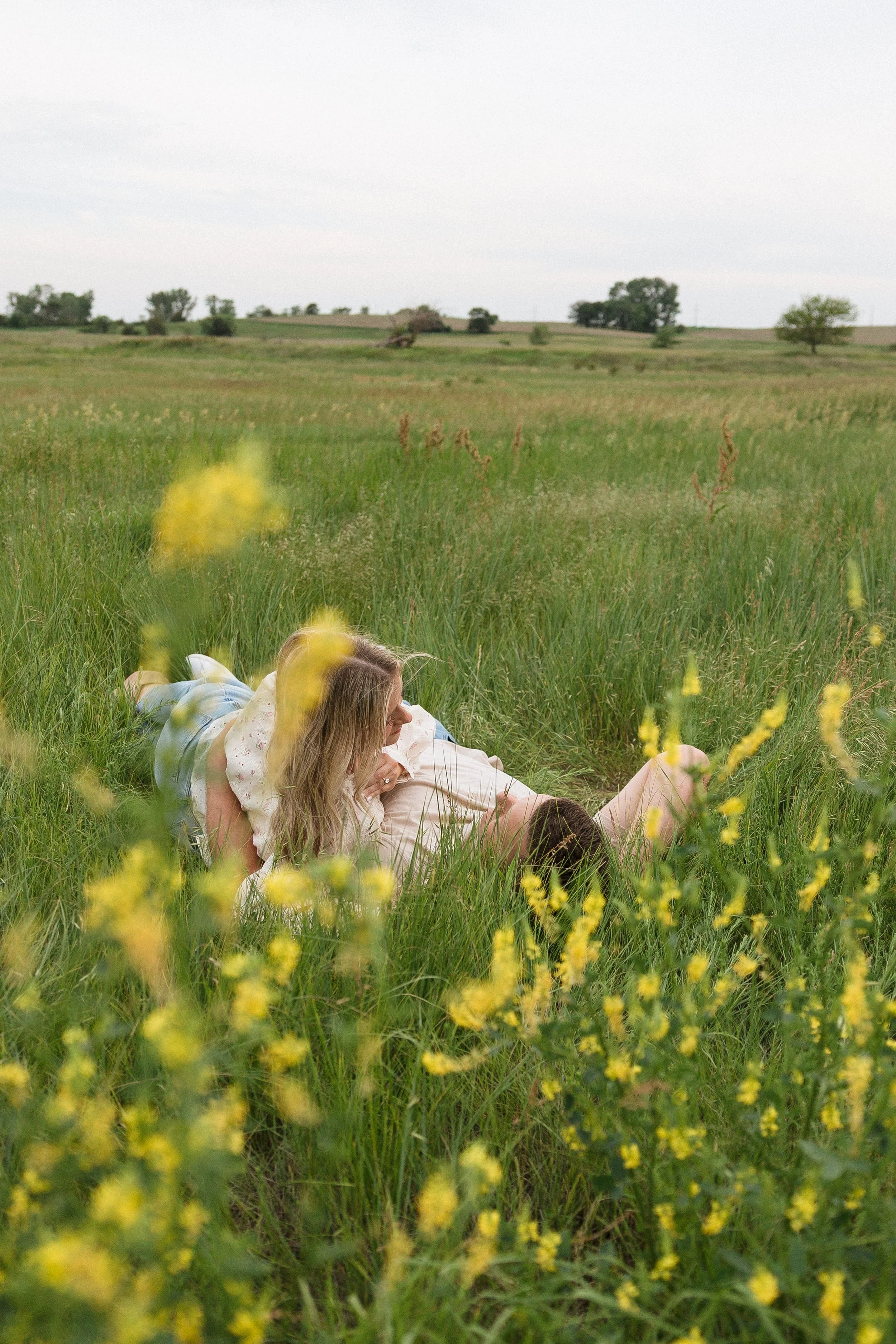 A couple lying in a grassy field with yellow wildflowers, embracing and looking at each other, with trees and open landscape in the background.