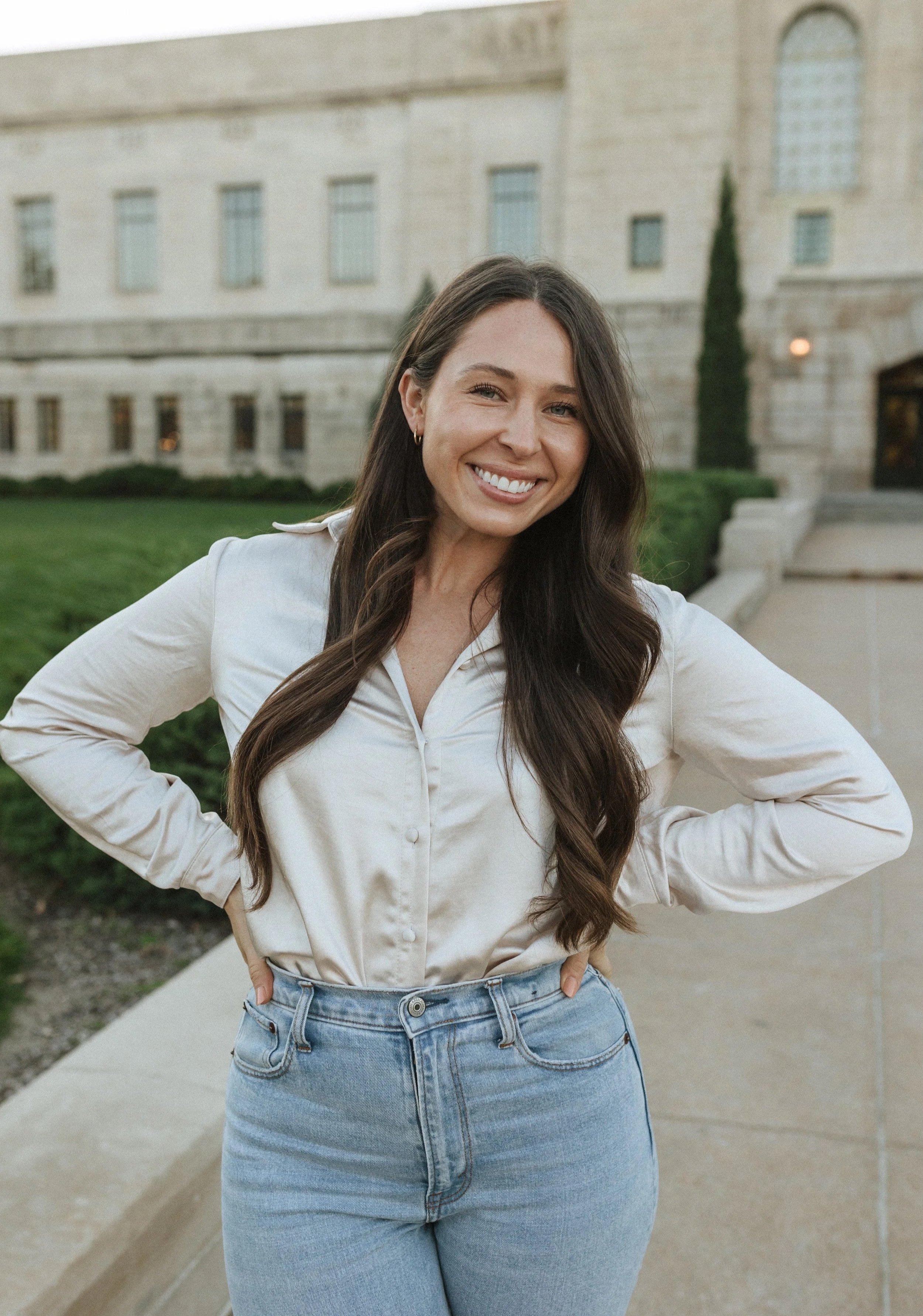 A woman with long dark hair smiling with her hands on her hips outside in front of a stone building with green bushes.