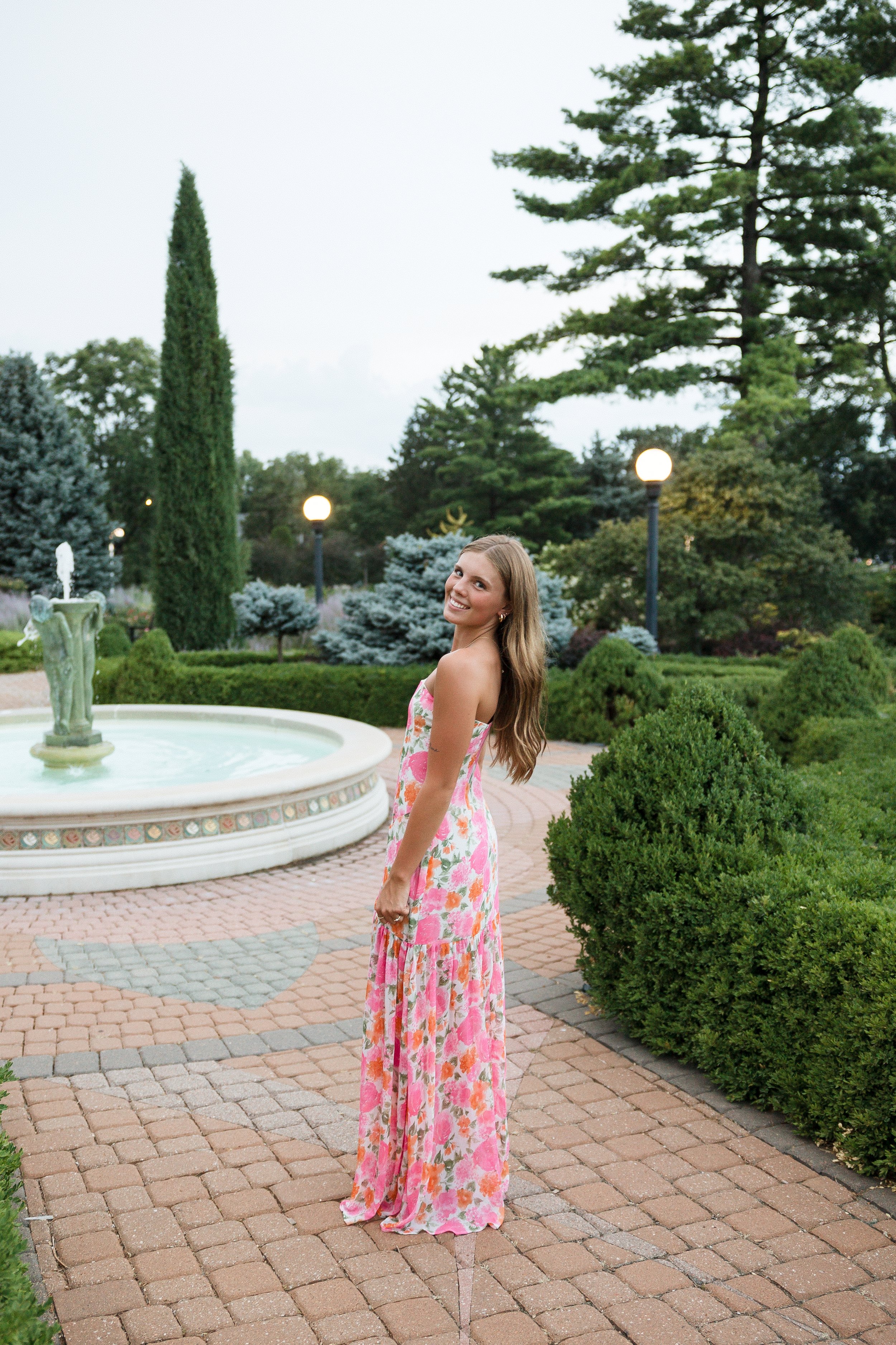 A woman in a floral maxi dress smiling in a garden with a fountain, trees, and lamp posts.