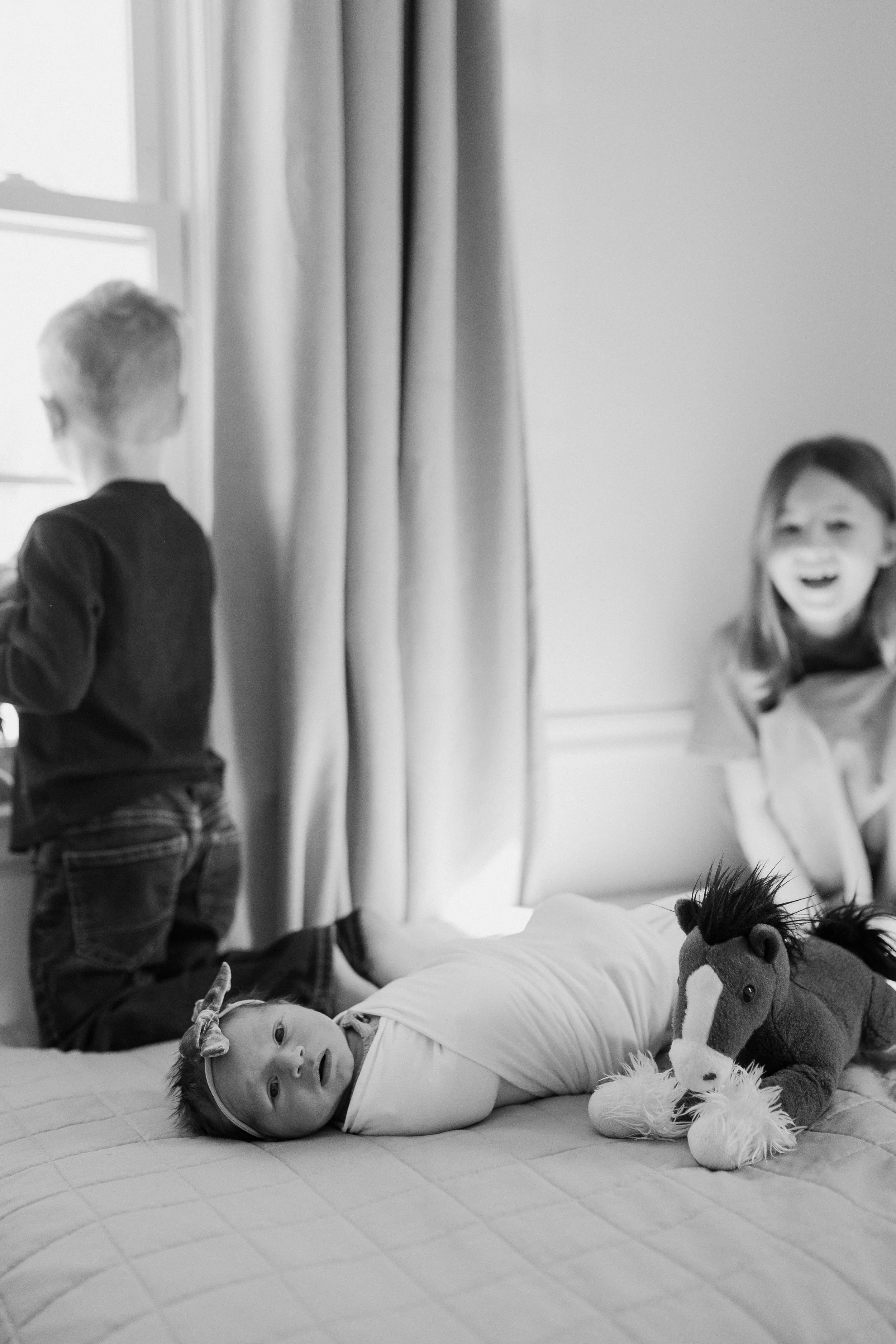 A baby lying on a bed with a stuffed horse toy, two children playing near a window, one girl smiling.