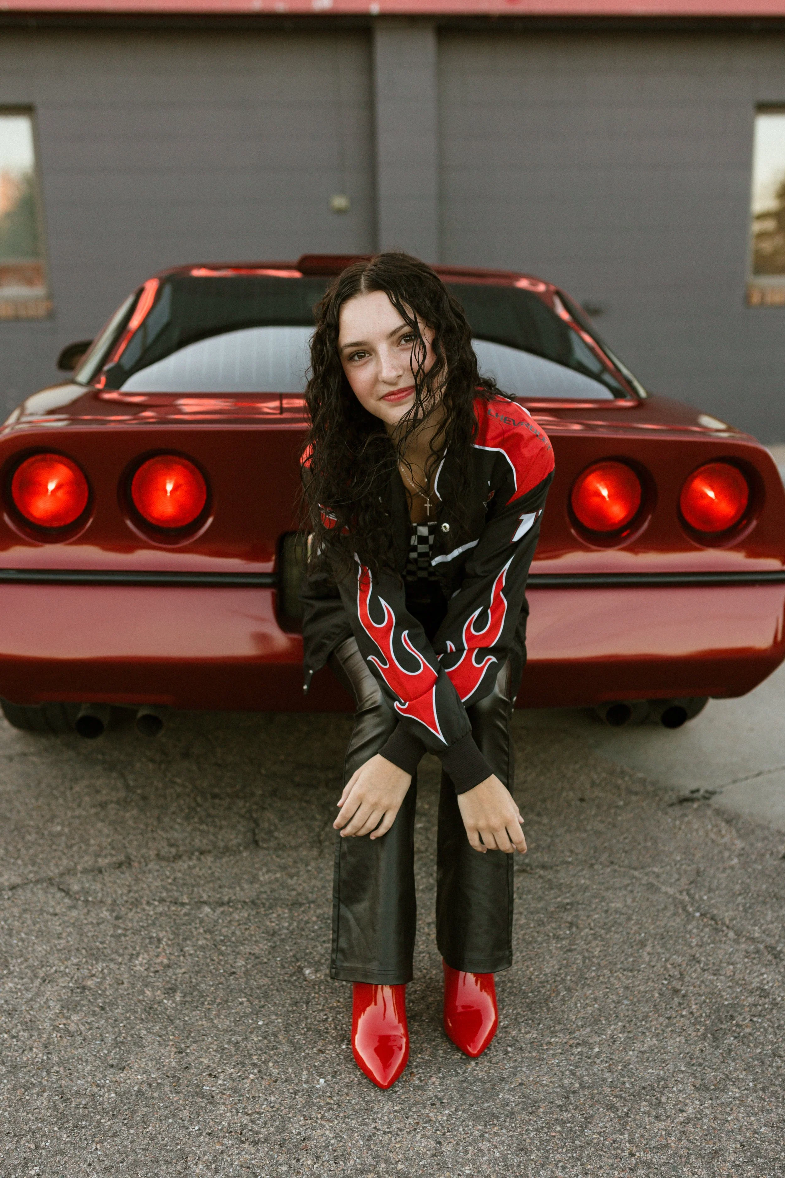 A young woman with curly dark hair sitting in front of a red sports car, wearing a racing jacket and high-heeled red boots.