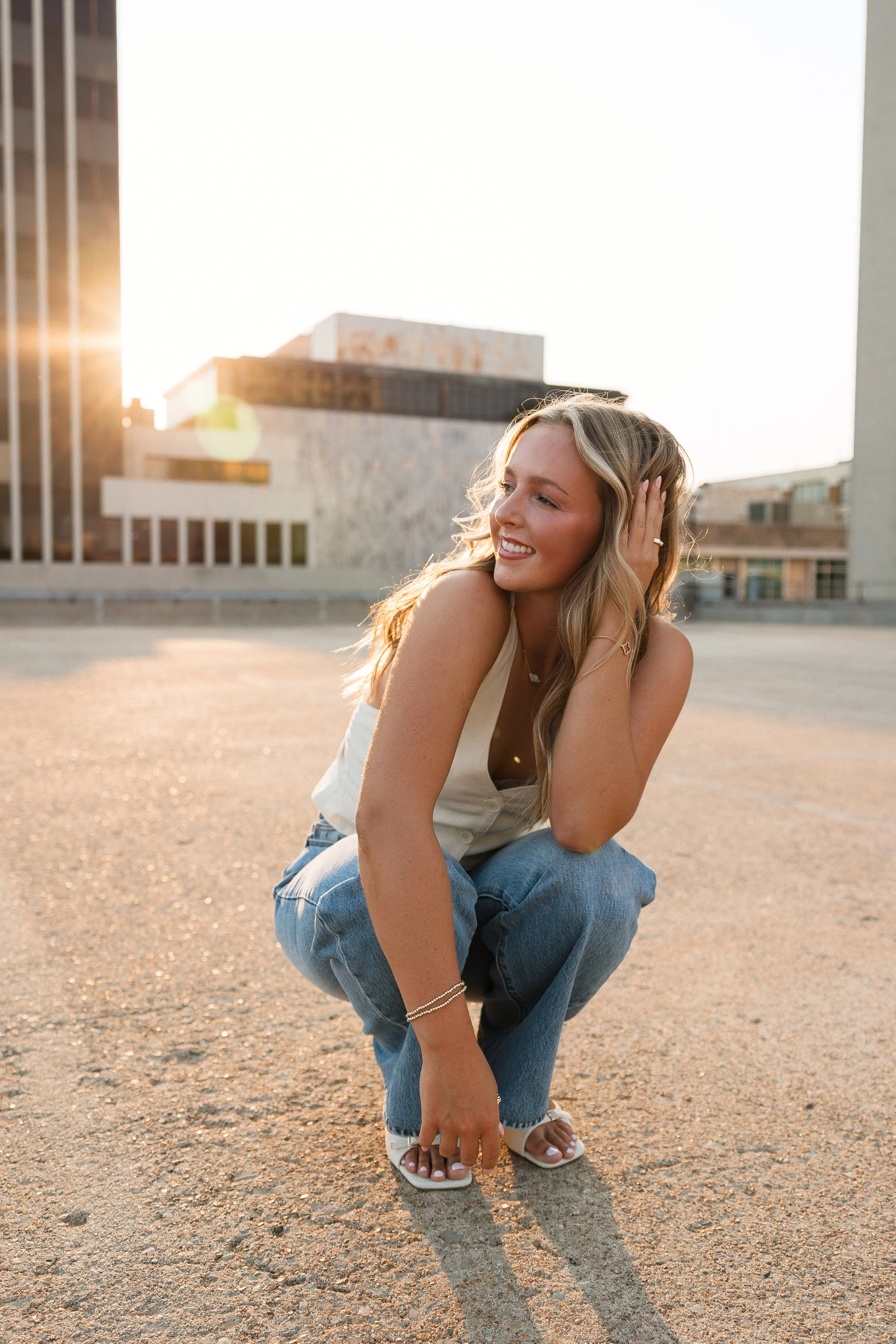 A young woman with long blonde hair smiling while crouching on a rooftop parking lot during sunset. She is wearing a white sleeveless top, blue jeans, and white sandals, with city buildings in the background.