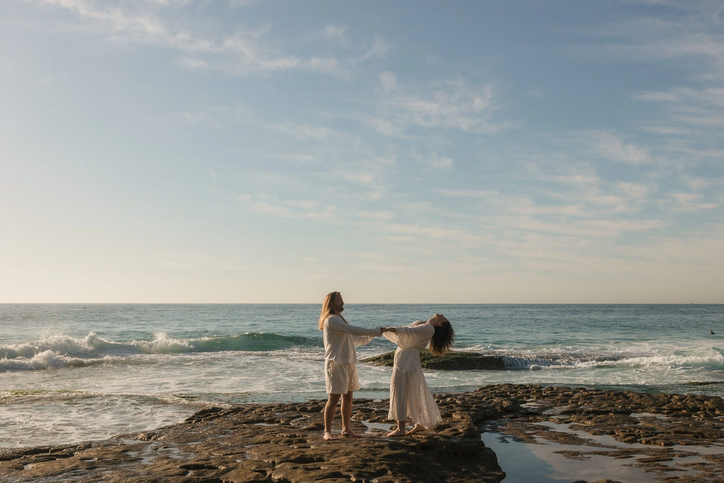 A couple standing on rocks by the ocean during sunset, holding hands and leaning back.