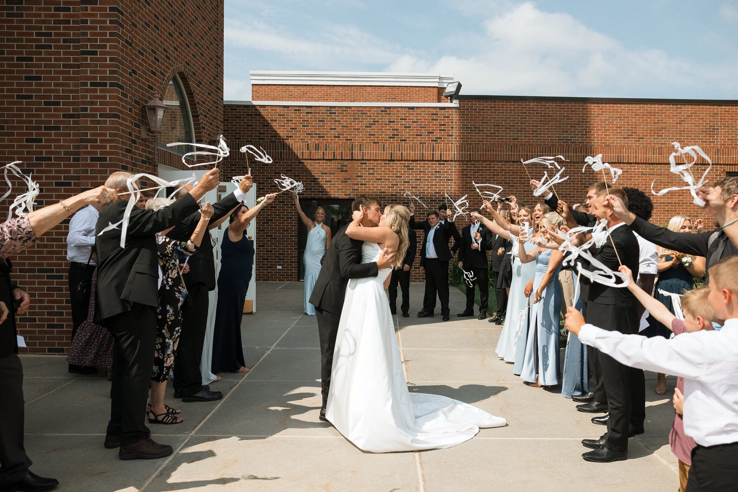 A bride and groom kissing in the center while friends and family celebrating around them, throwing white ribbon streamers in the air outside a brick building.