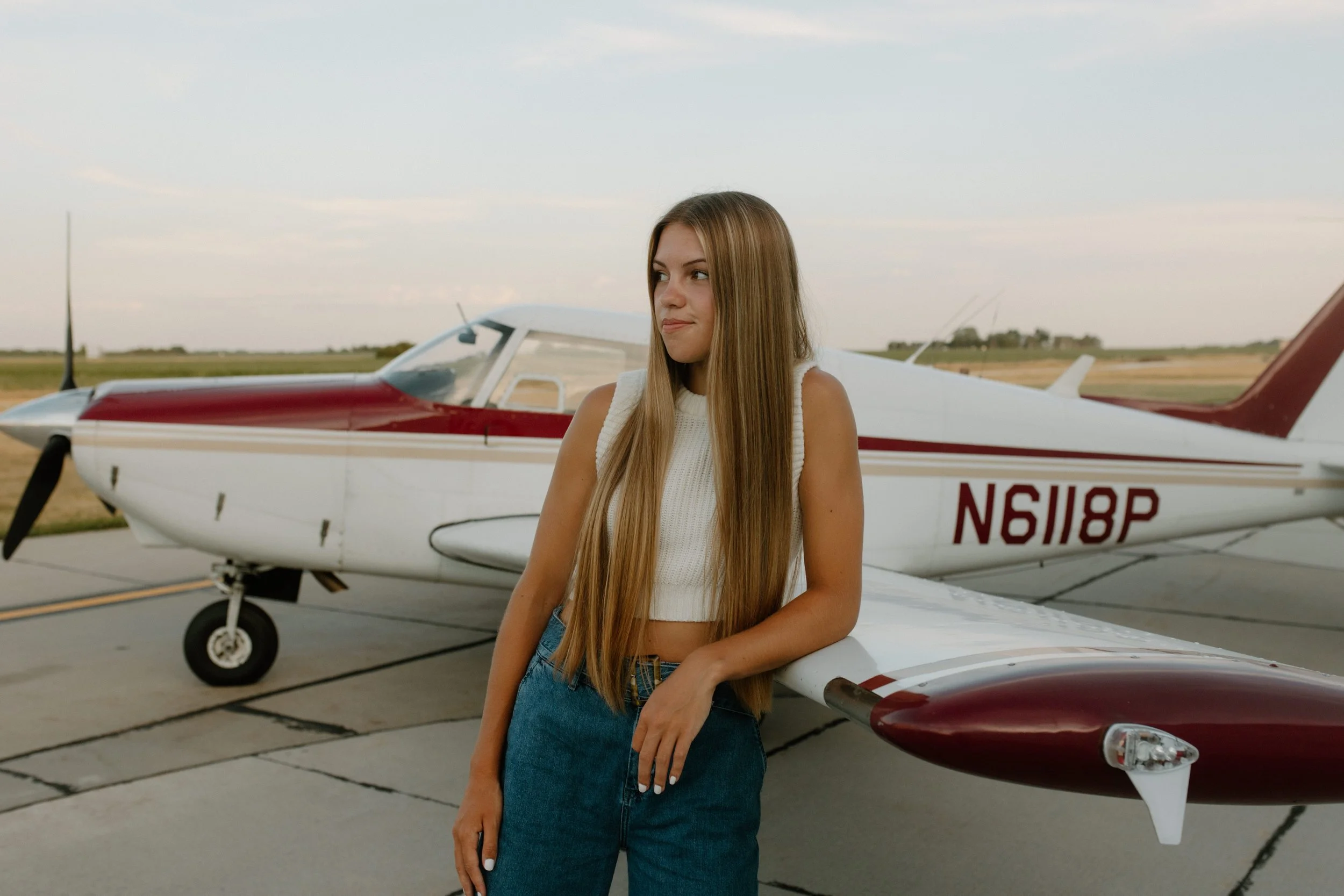 A young woman with long, straight hair leaning on a white and burgundy small airplane on a tarmac at sunset.