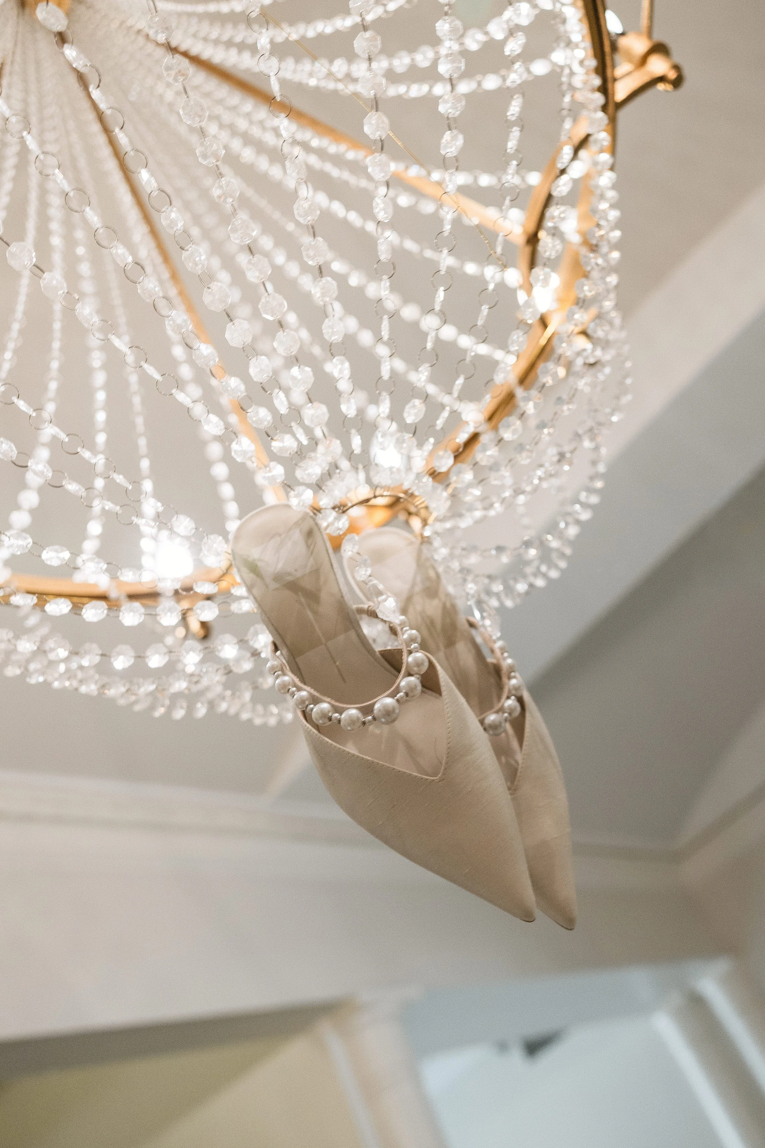 Beige high heel shoes hanging from a crystal chandelier with pearl decoration.