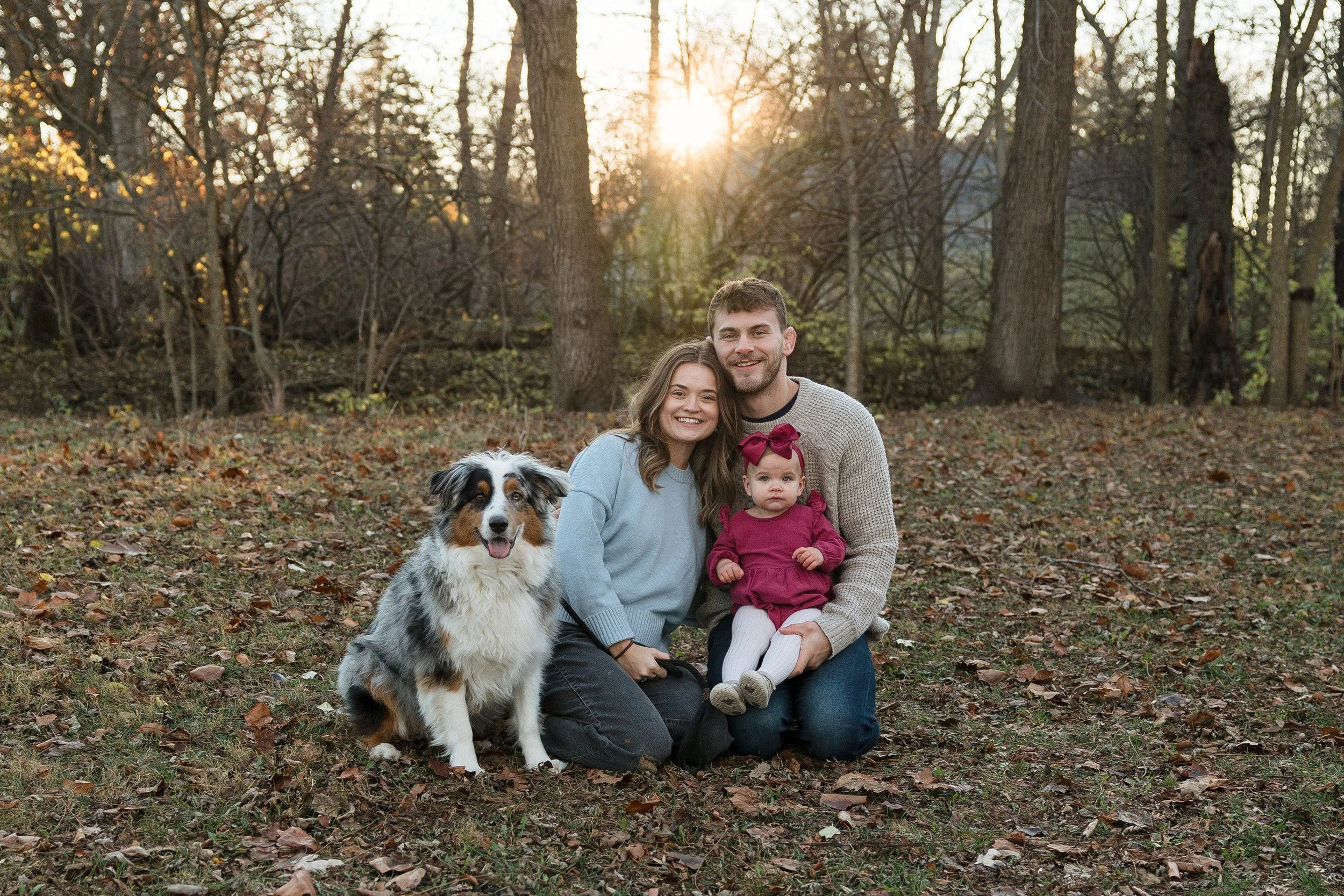A family of three with a dog sitting on fallen leaves in a wooded area during sunset, smiling at the camera.
