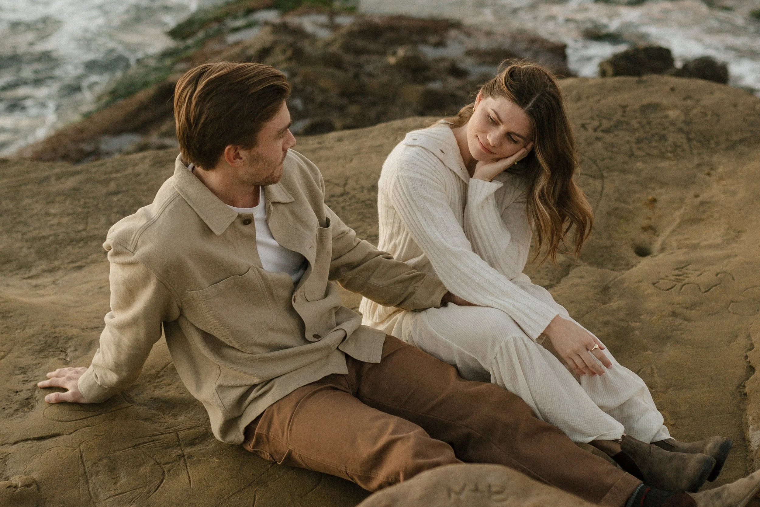 A man and woman sitting on a sandy beach near the rocky shoreline, with waves in the background. The woman is resting her head on her hand and looking at the man, who is leaning back on one arm and looking at her.