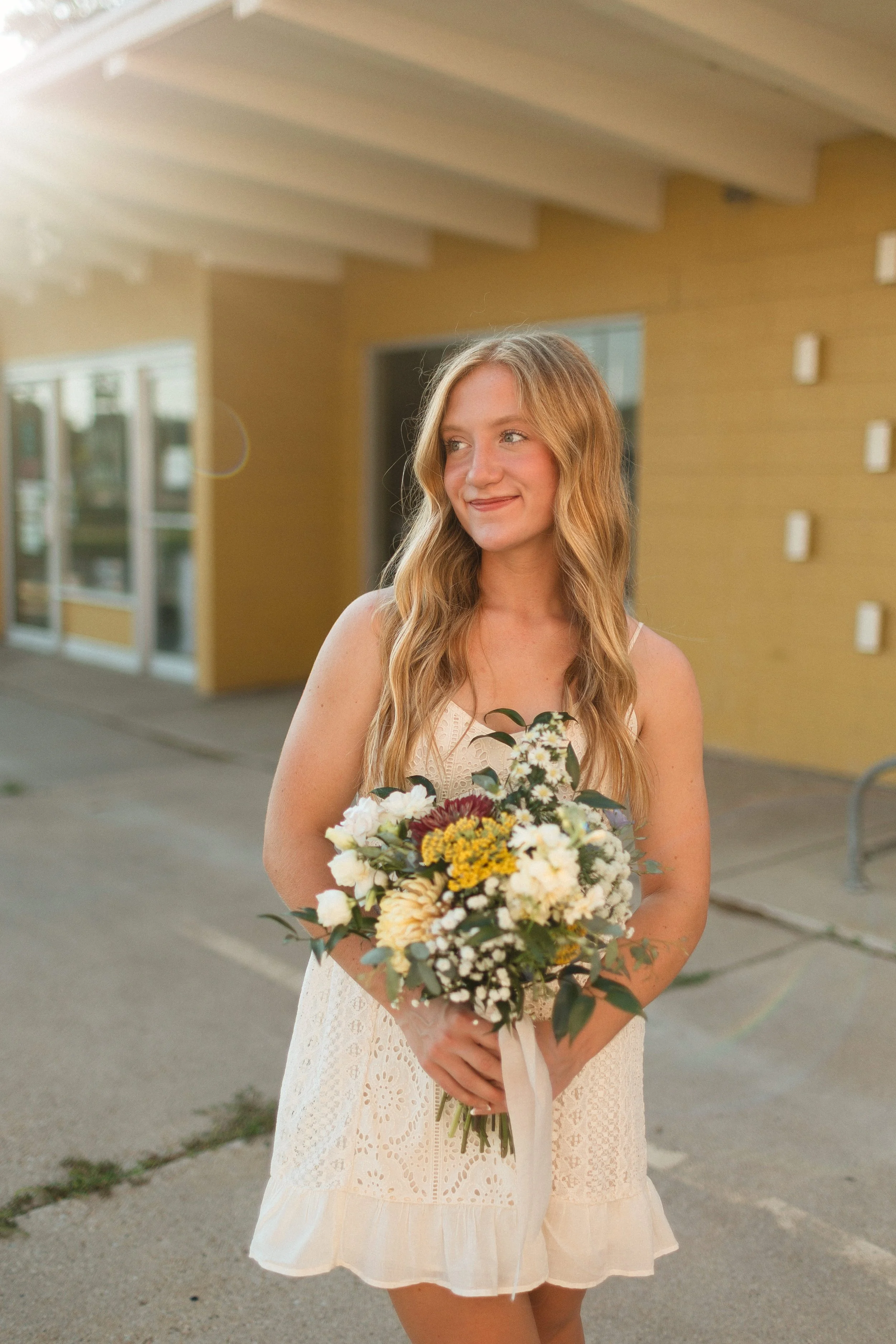 A young woman with long wavy blonde hair, wearing a sleeveless cream-colored dress, holding a bouquet of flowers, standing outside near a yellow building at sunset.