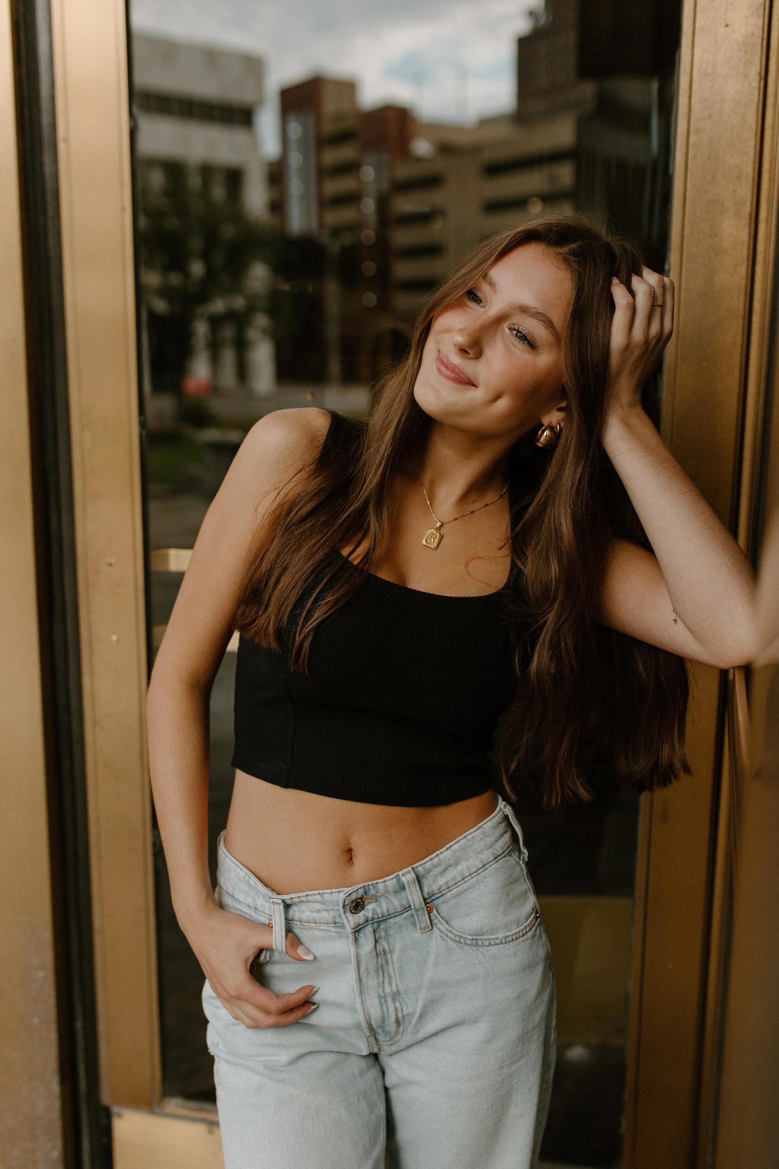 A young woman with long brown hair wearing a black crop top and light blue jeans posing inside near a glass door with an urban cityscape in the background.
