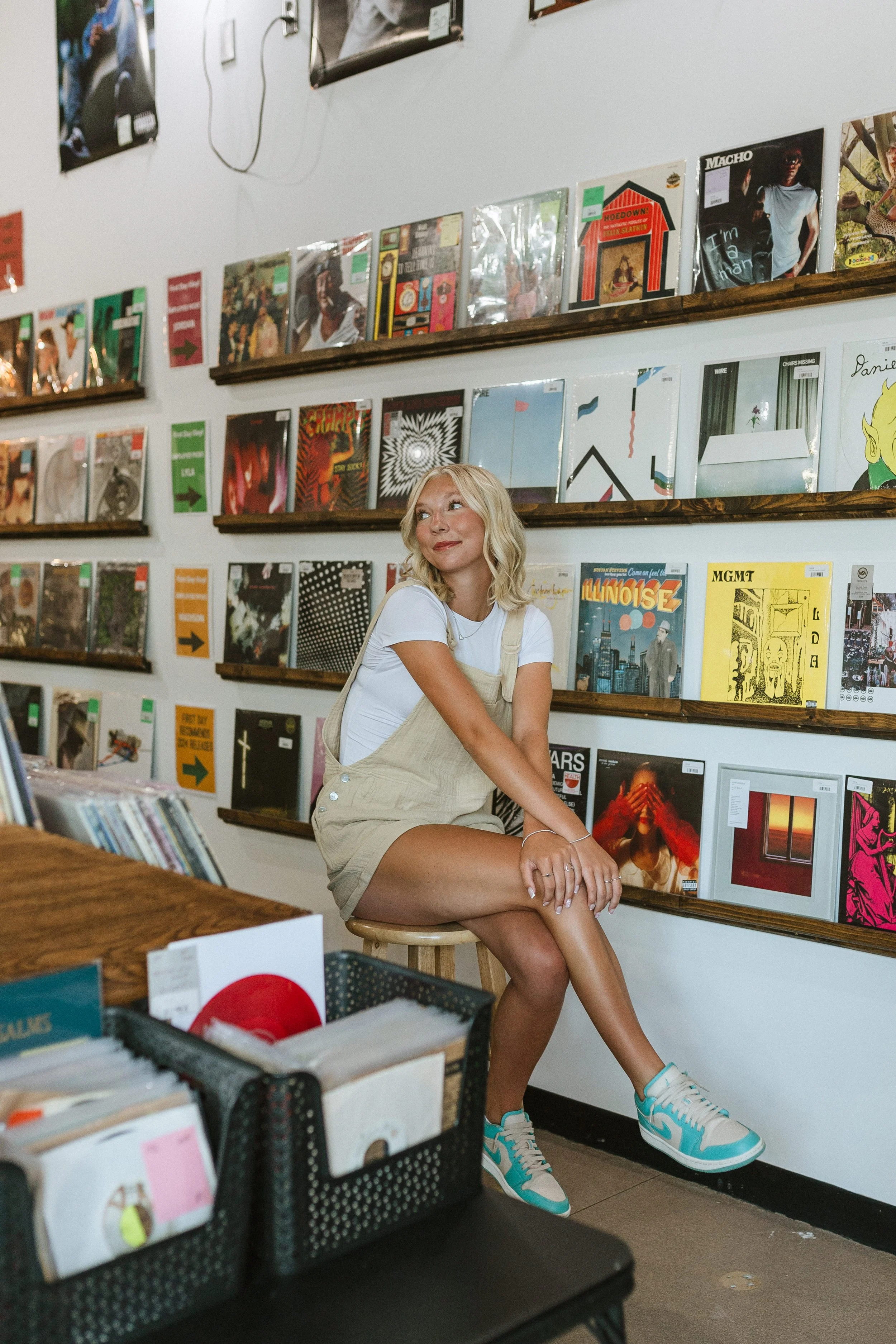 A young woman with blonde hair sitting on a stool in a record shop, surrounded by shelves of vinyl records.