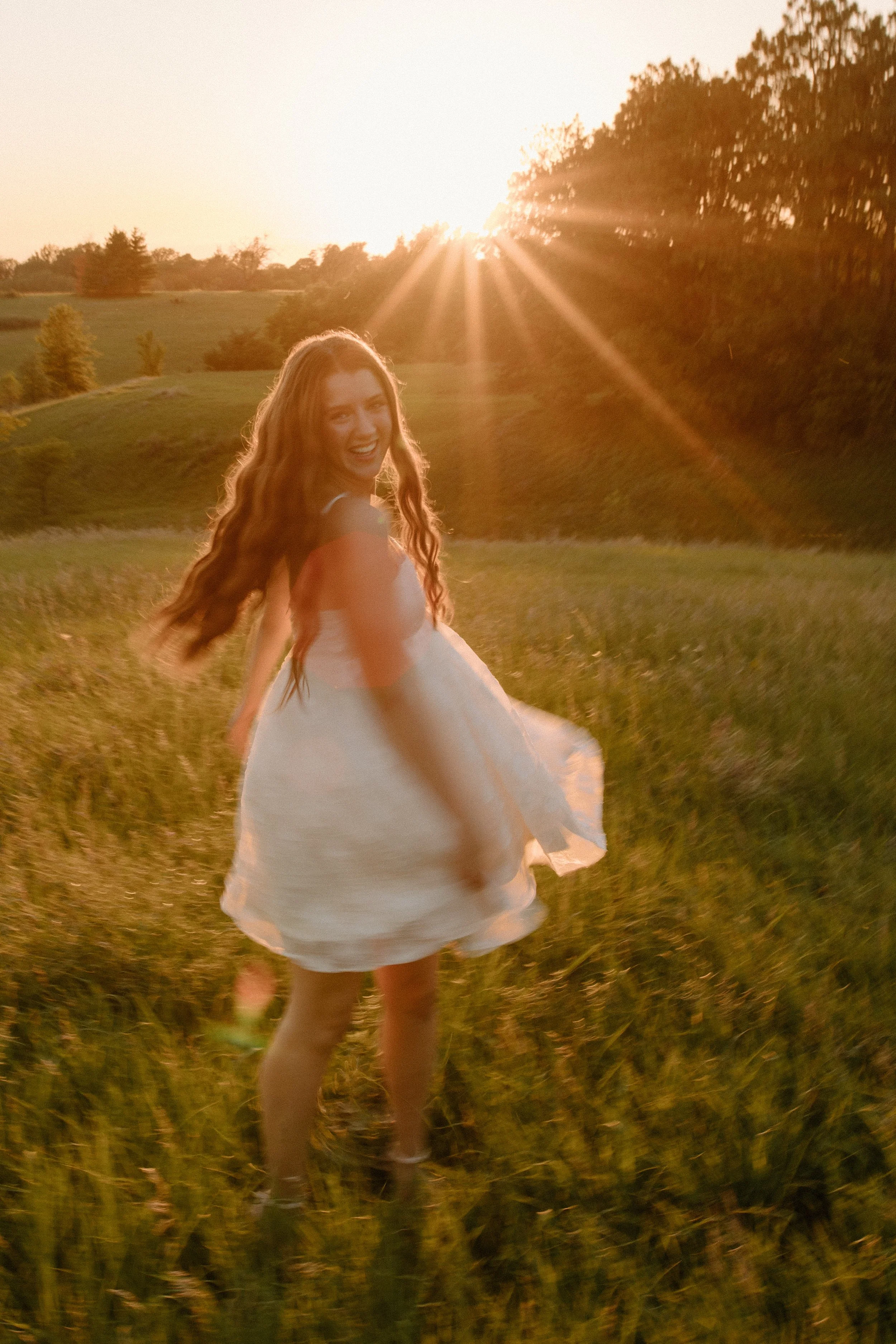 A girl with long wavy hair wearing a white dress standing in a grassy field during sunset, smiling and looking back at the camera with the sun shining in the background.