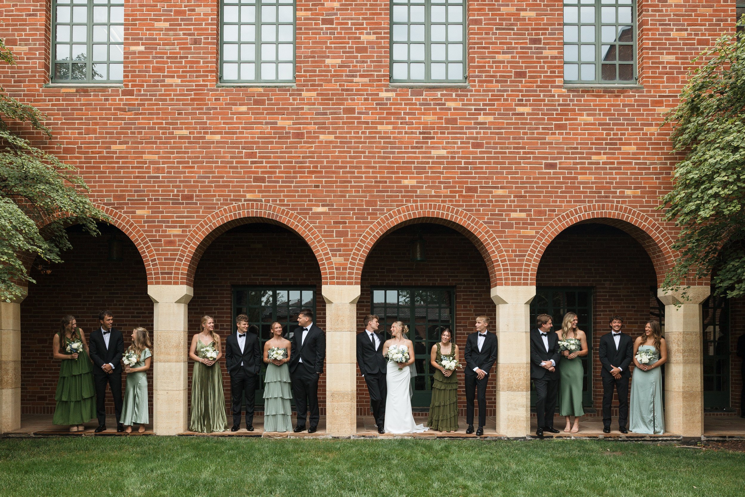 Wedding party standing under brick arches, with the bride and groom in the center, surrounded by bridesmaids in green dresses and groomsmen in black tuxedos.