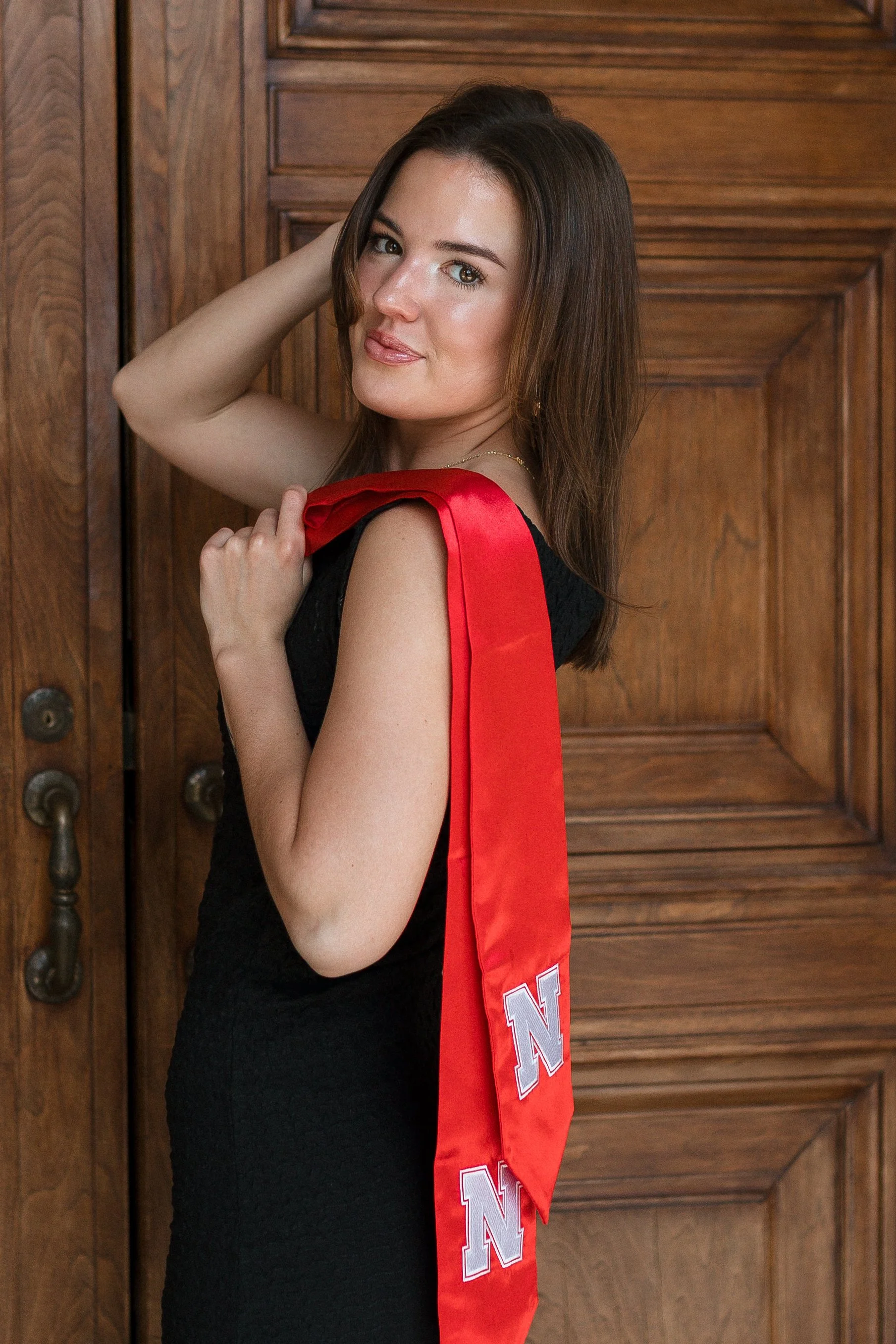 A young woman with long brown hair, wearing a black dress and an 'N' lettered red sash, stands in front of a wooden door, smiling slightly and looking at the camera.