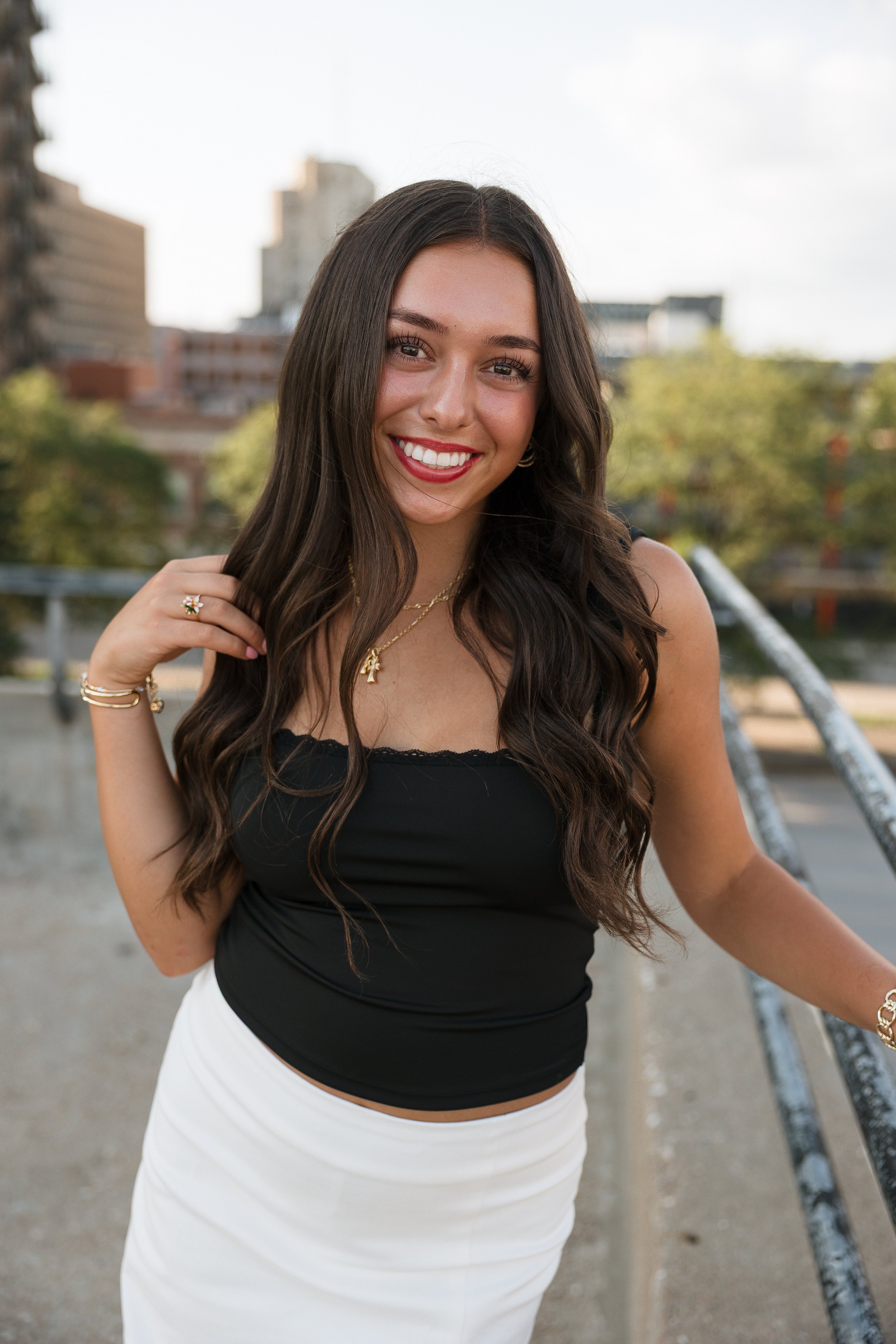 A young woman with long dark hair and a bright smile, wearing a black sleeveless top and a white skirt, standing outdoors on a bridge with city buildings and trees blurred in the background.