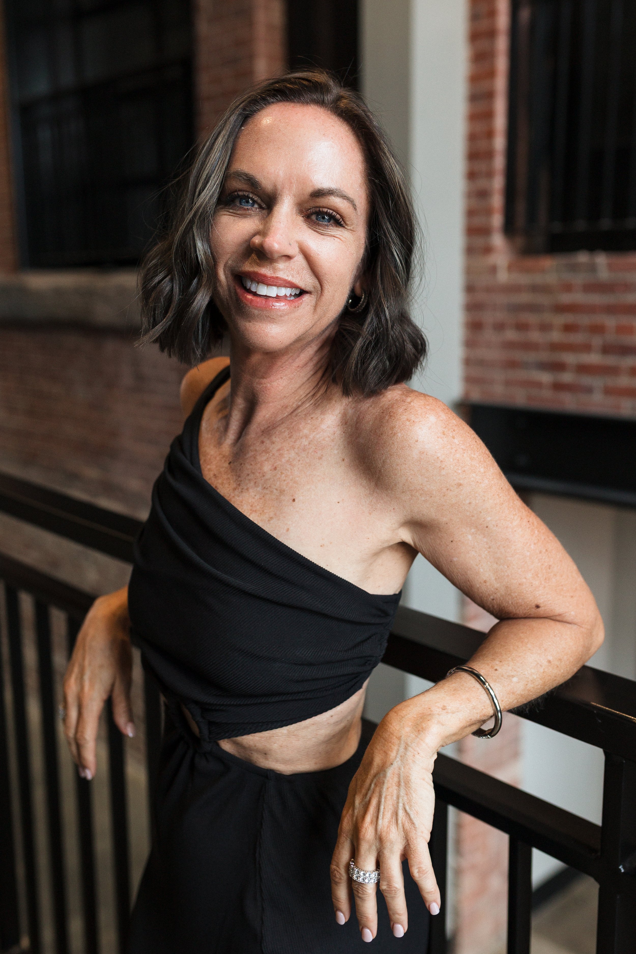 A woman with short brown hair, wearing a black one-shoulder top, smiling, leaning on a black railing near a brick wall.