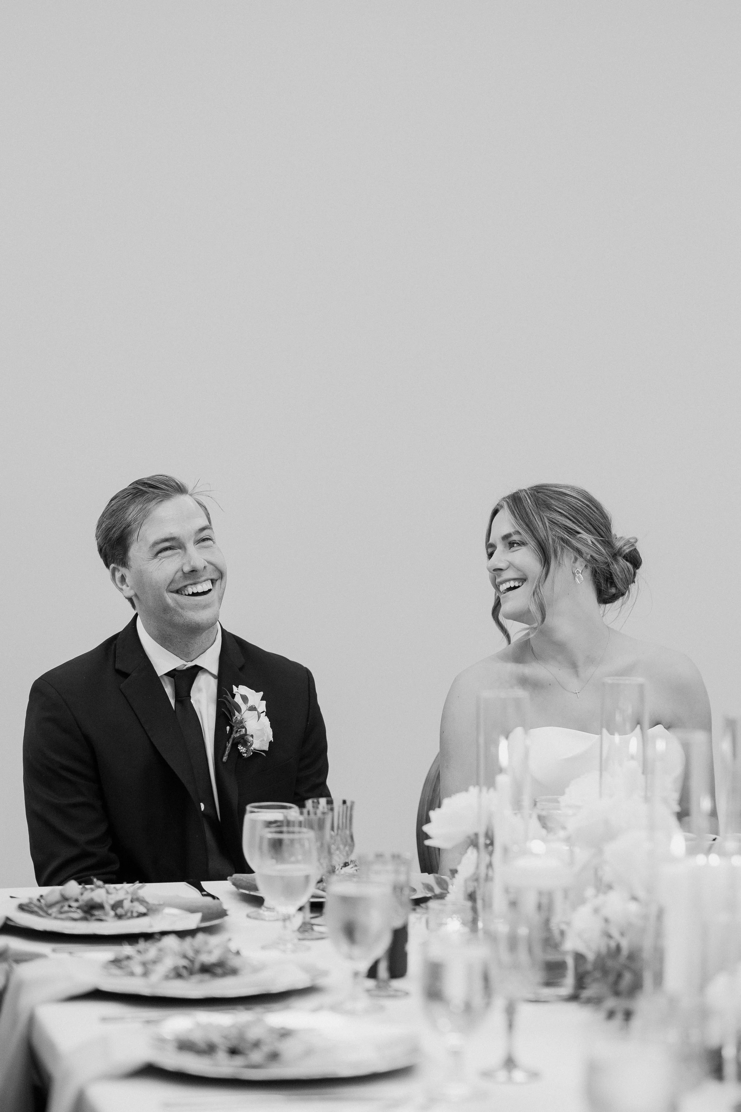 A black and white photo of a smiling man and woman sitting at a reception table, dressed in formal attire, likely at a wedding.