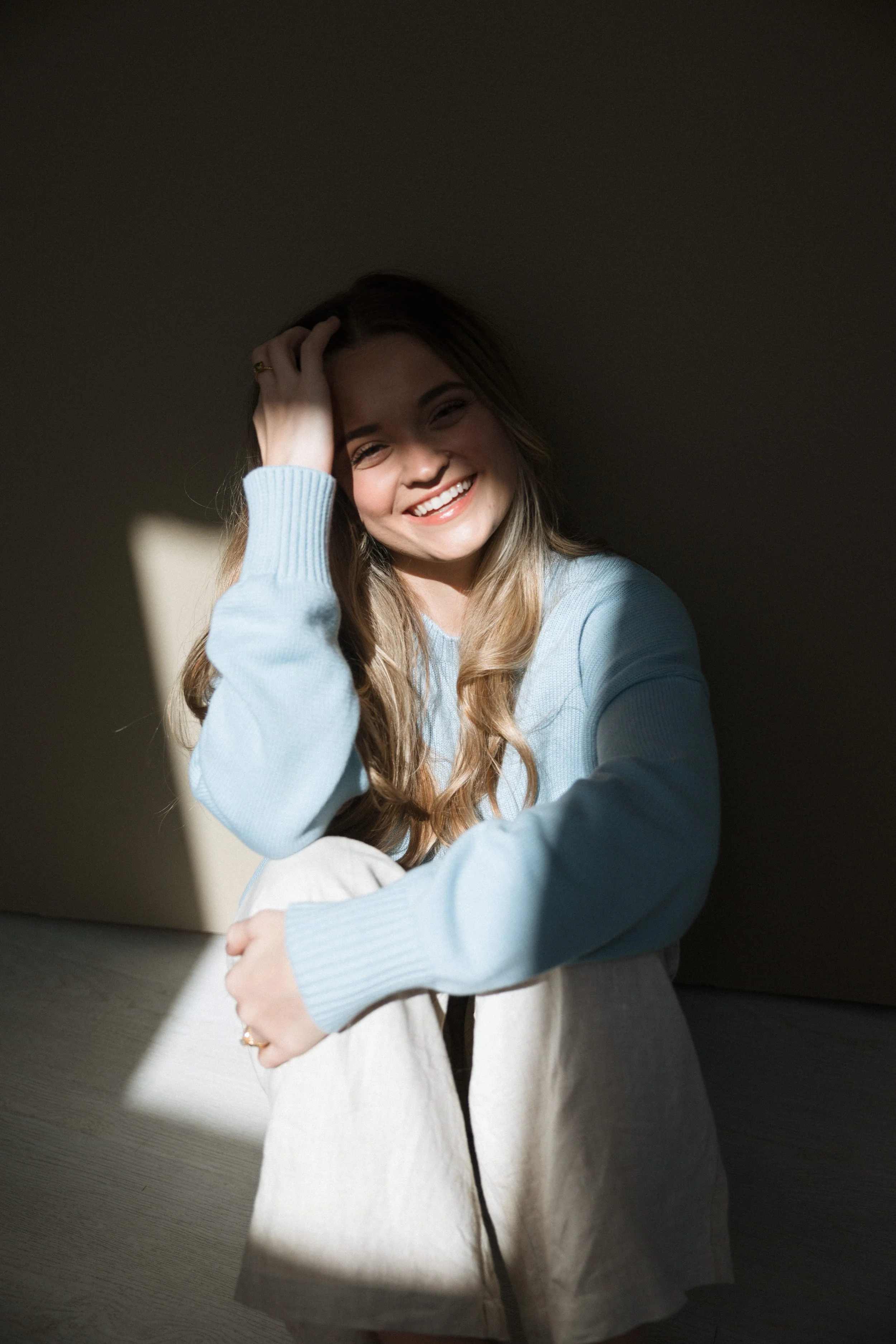 A young woman with long, wavy hair smiling and laughing, sitting with her legs crossed in a space with partial sunlight and shadow on her face and body.