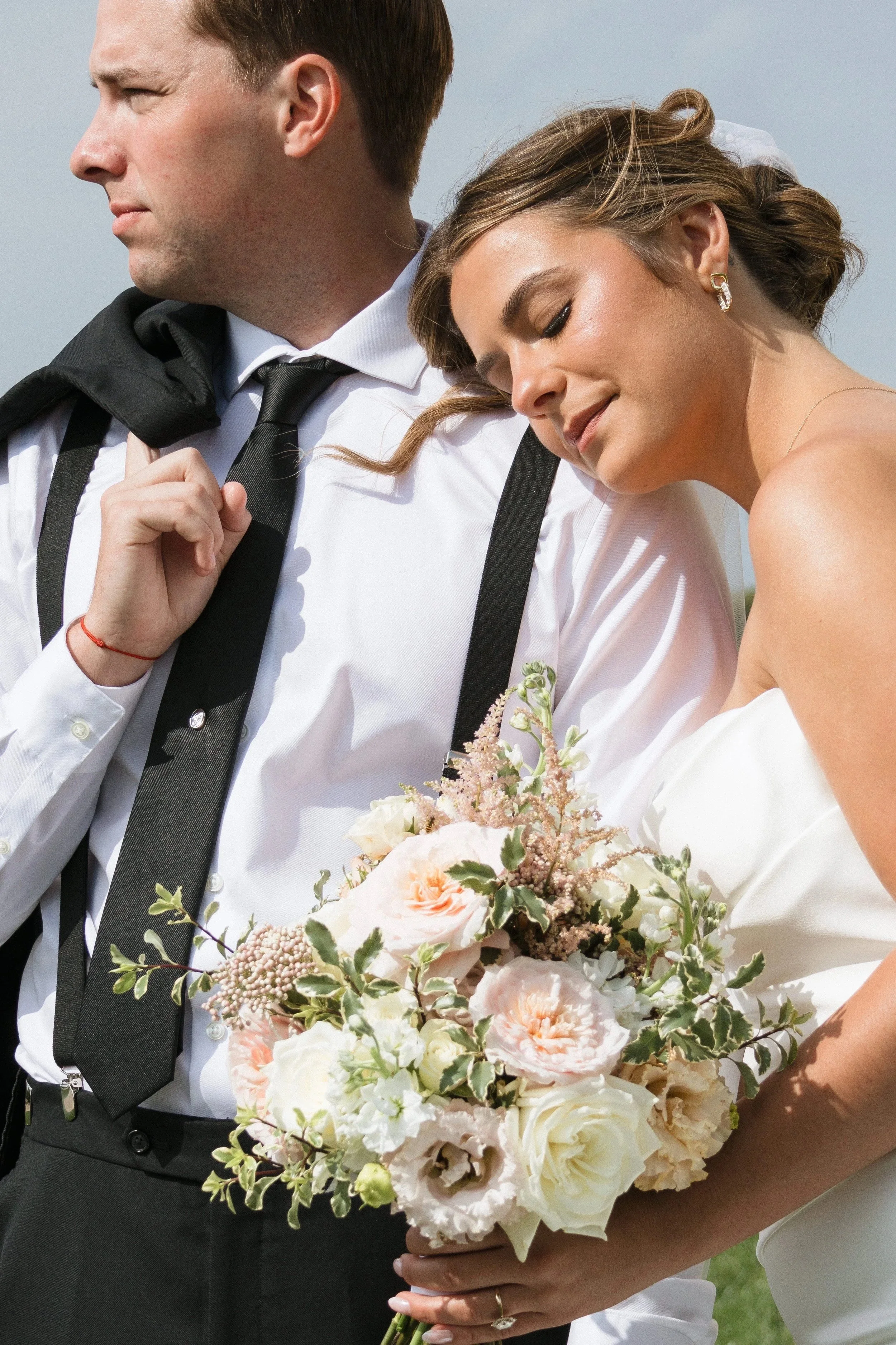 A bride and groom embrace outdoors, the bride holding a bouquet of pink and white flowers, both dressed in wedding attire.
