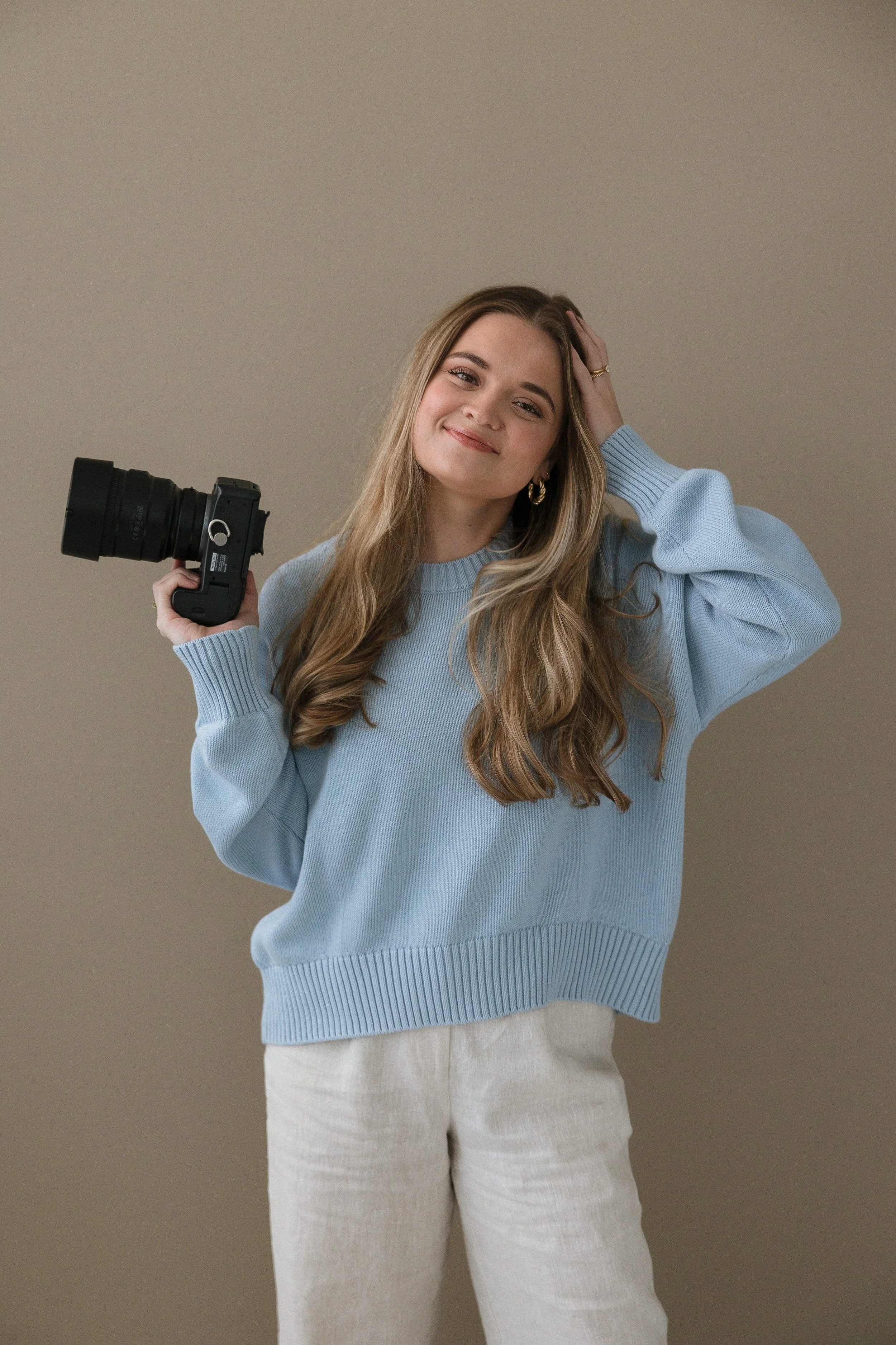A smiling young woman with long wavy hair, wearing a light blue sweater and beige pants, holding a camera in her right hand against a plain beige background.