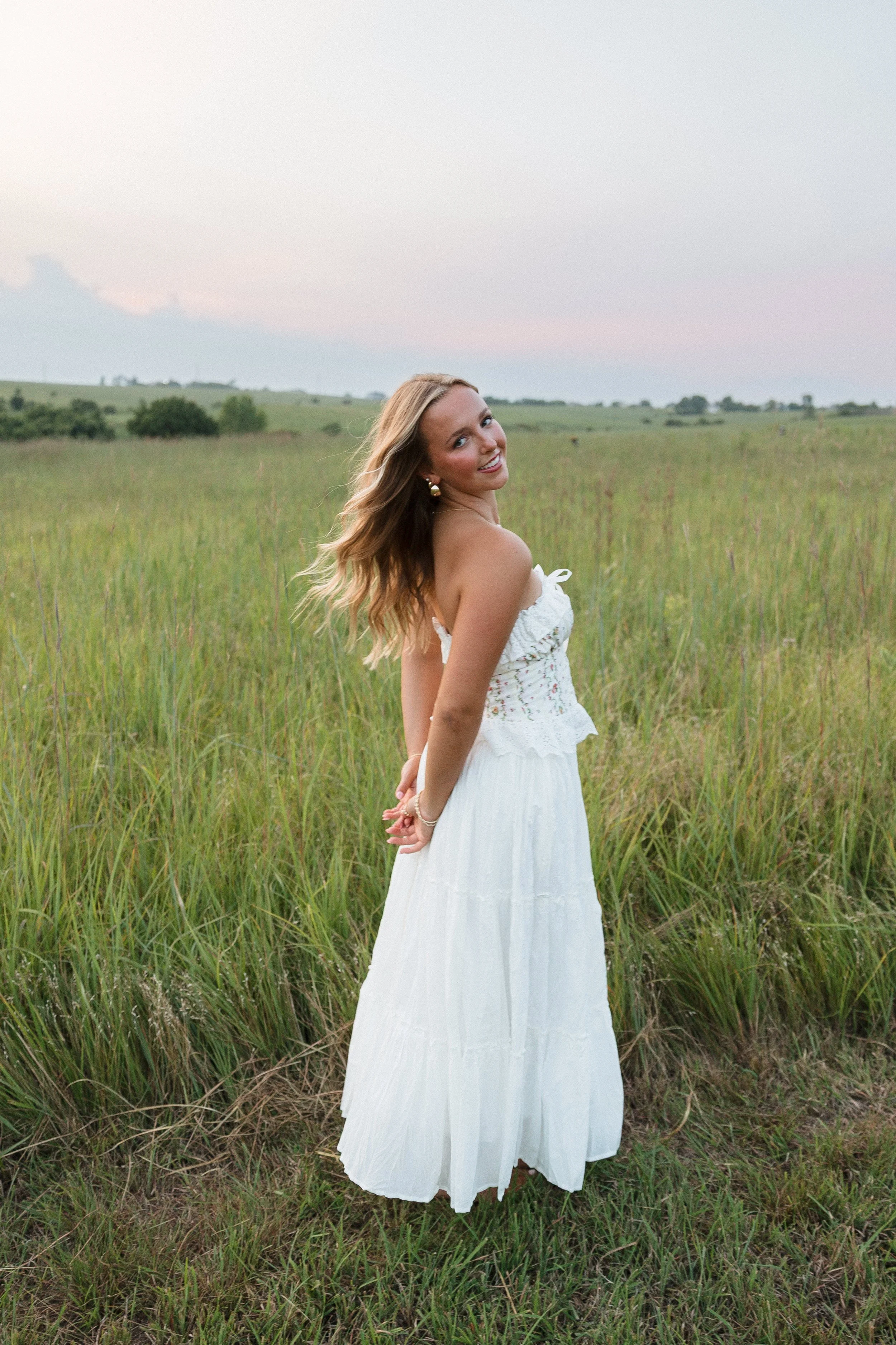Woman in a white dress standing in a grassy field at sunset, smiling and looking back.