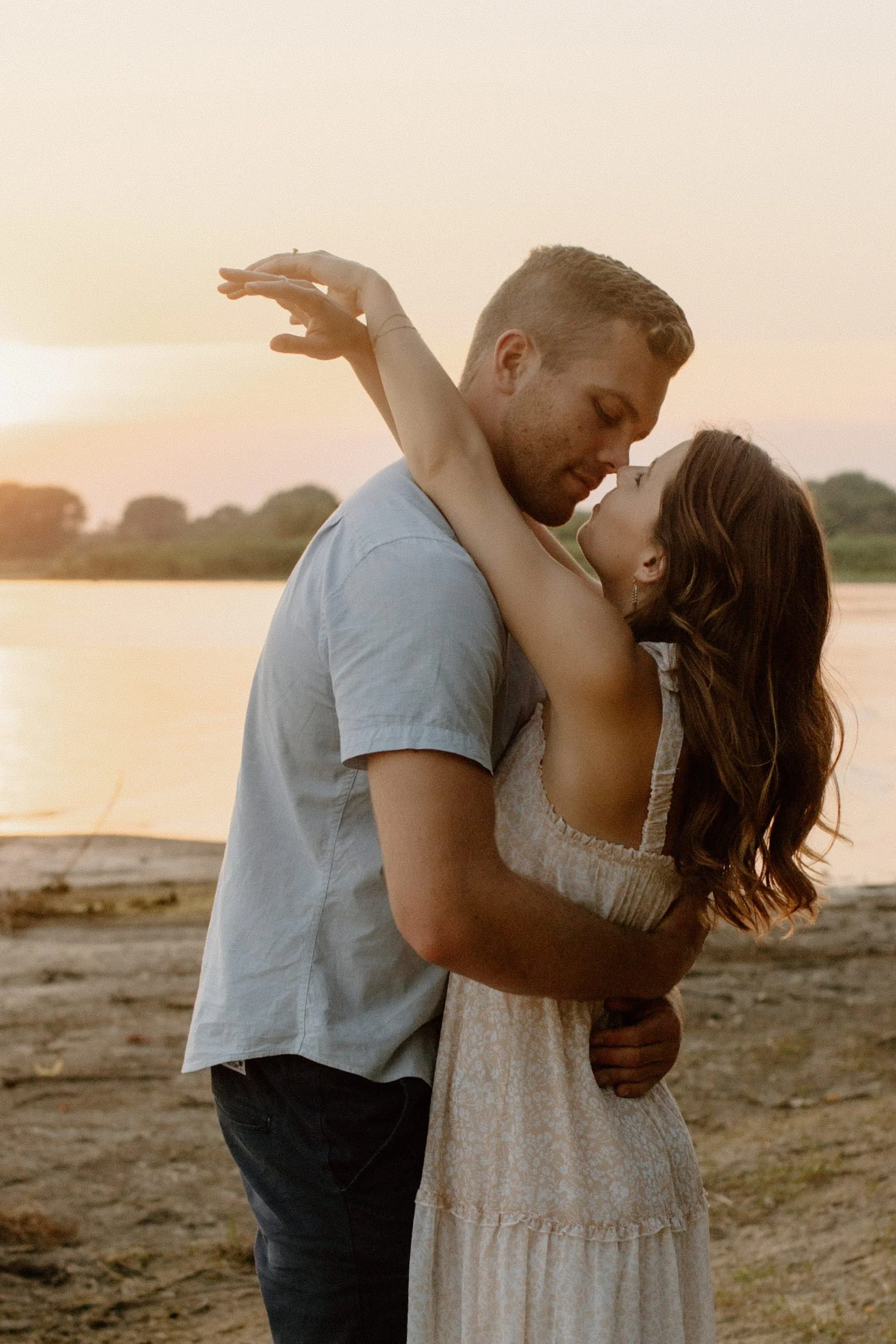 A couple sharing a romantic embrace by a river during sunset, looking into each other's eyes.