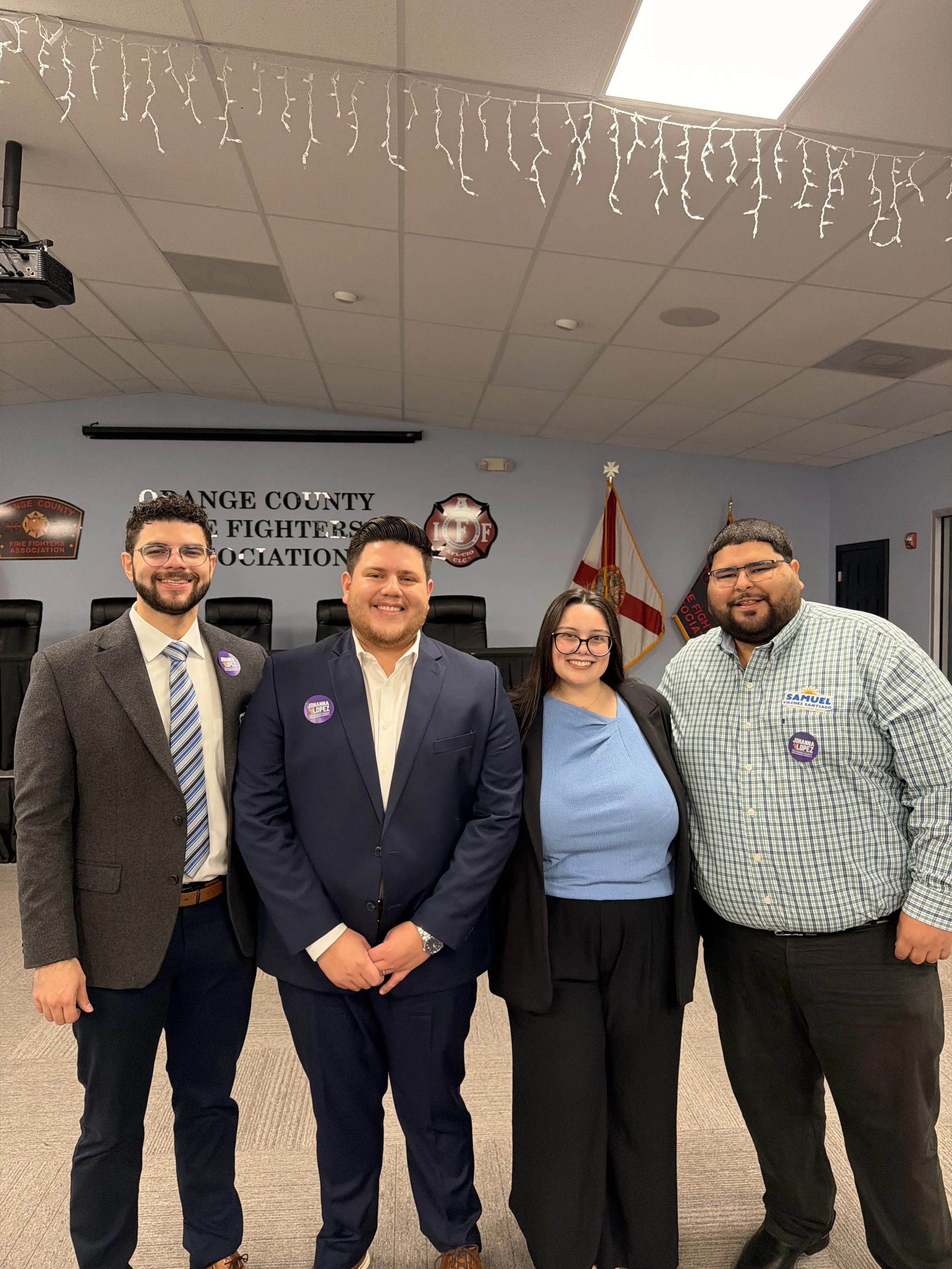 Four people standing together in a meeting room with a sign behind them that reads 'Orange County Fire Fighters Association,' a fire department badge, and Florida state flags.