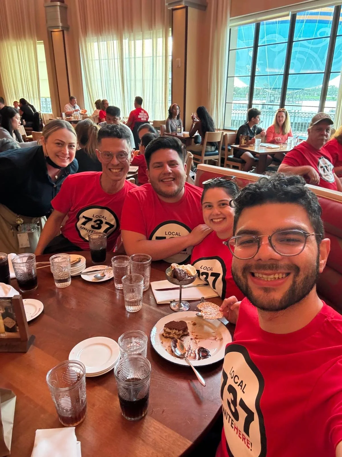 A group of five young adults smiling at a restaurant table with desserts, drinks, and plates, wearing matching red shirts with a black and white 'Local 737' logo.