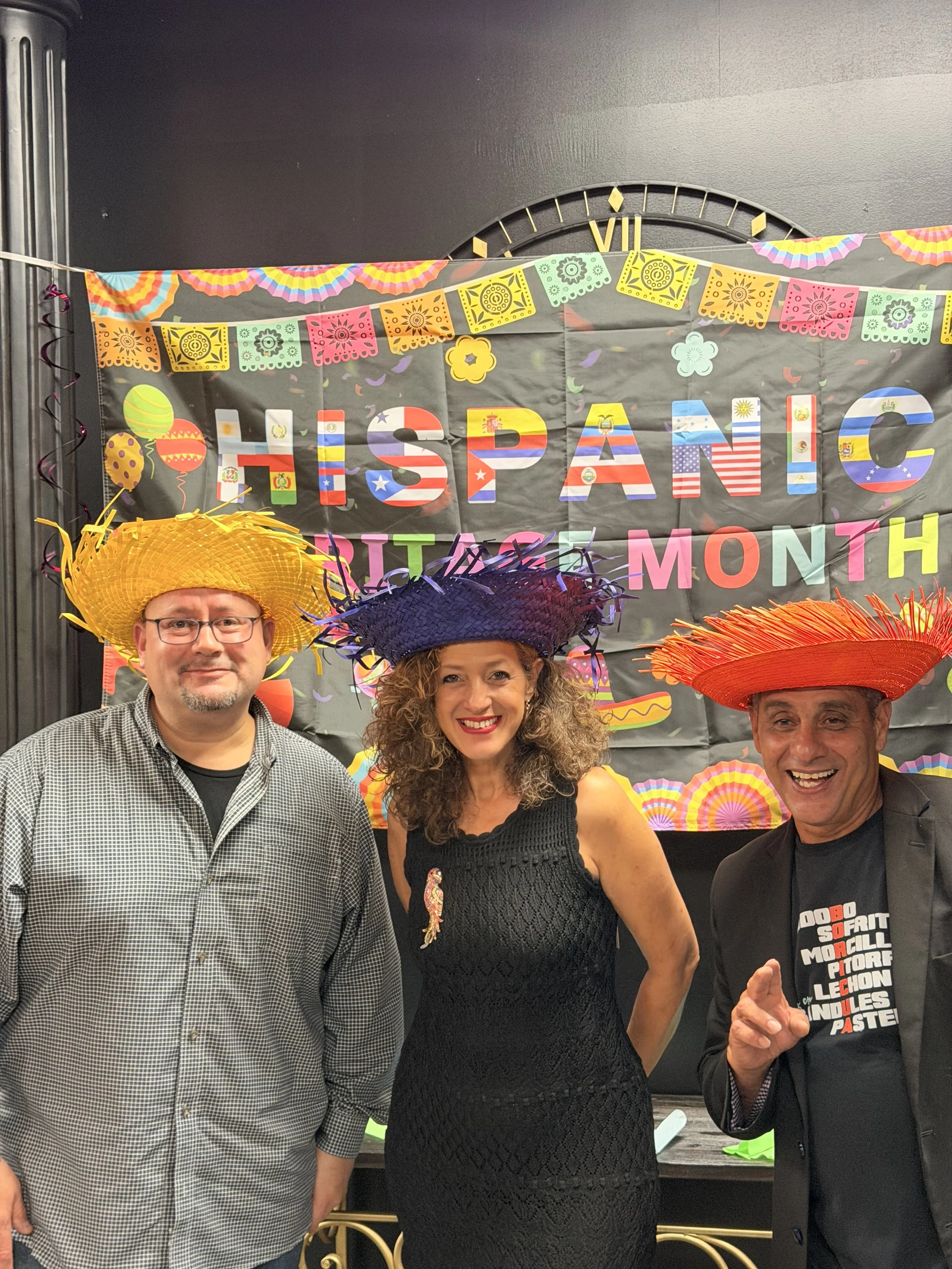 Three people wearing colorful sombreros, standing in front of a Spanish National Day celebration poster.