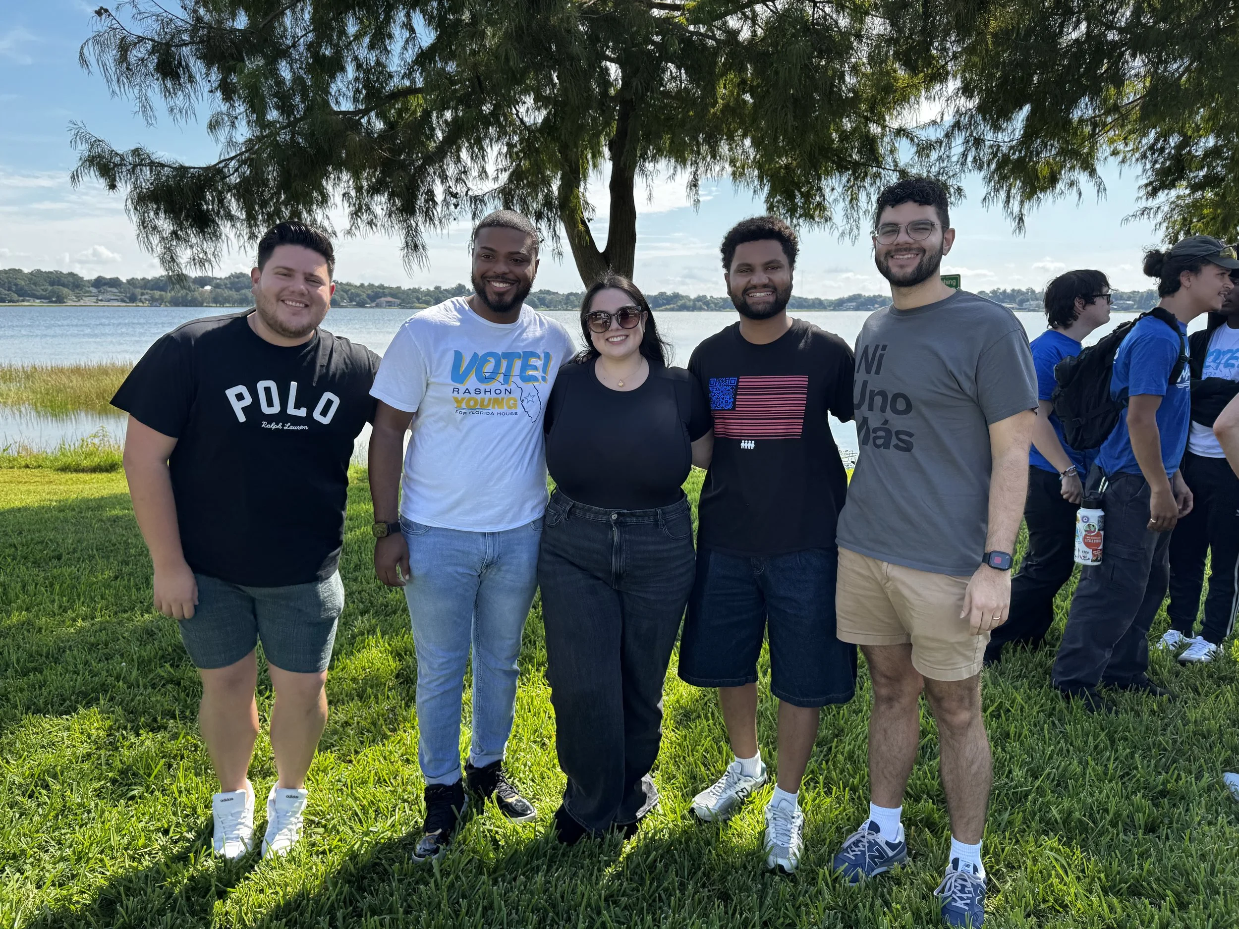Group of six people standing outdoors on grass near a body of water with trees and blue sky in the background. They are smiling and posing for the photo.