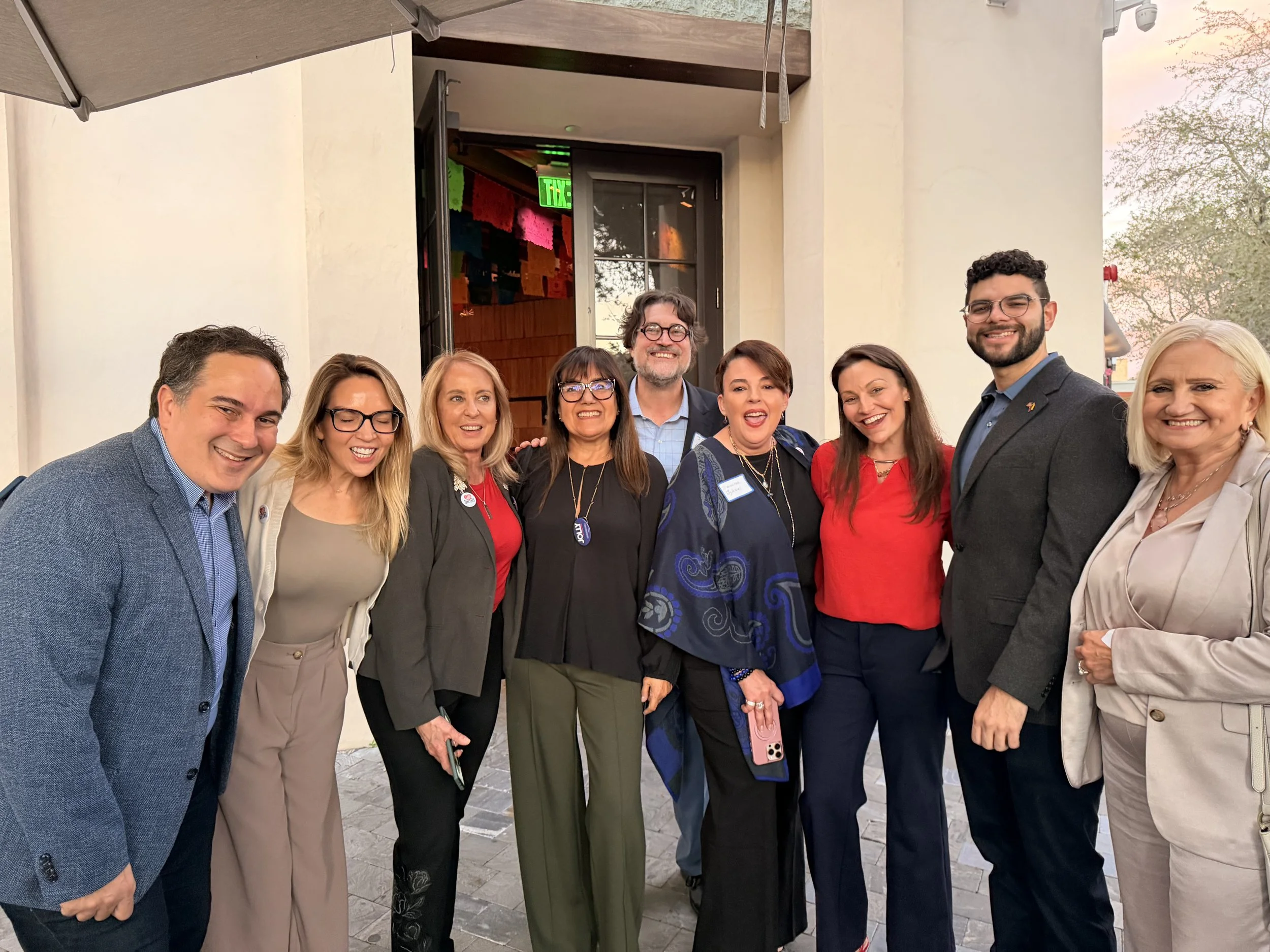 A group of ten people standing together outdoors, smiling, dressed in business casual attire, with a building entrance and some trees in the background.