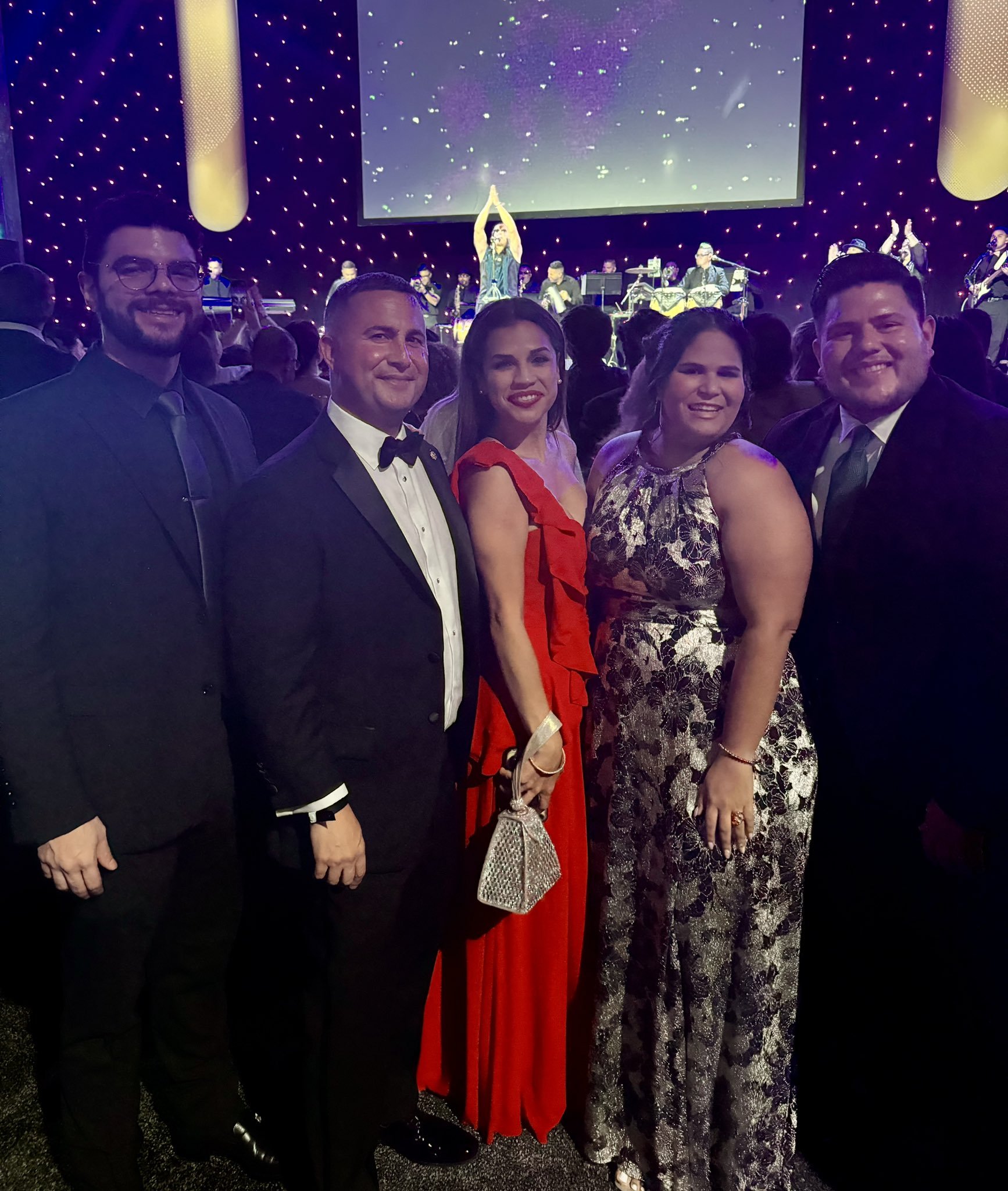 Group of five people dressed in formal attire posing at a concert or event with live music on stage in the background.