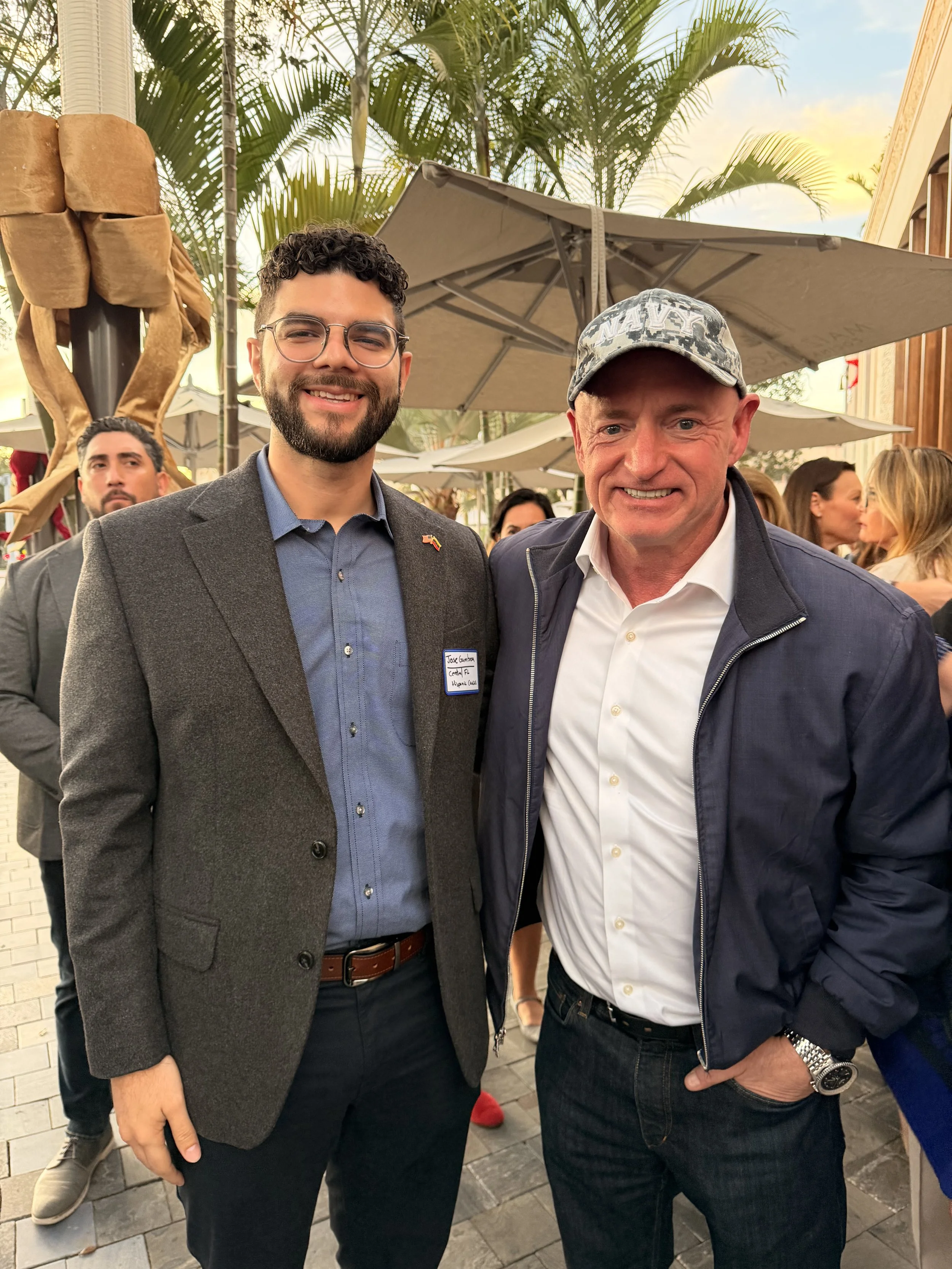 Two men smiling at an outdoor event, with palm trees and other people in the background.