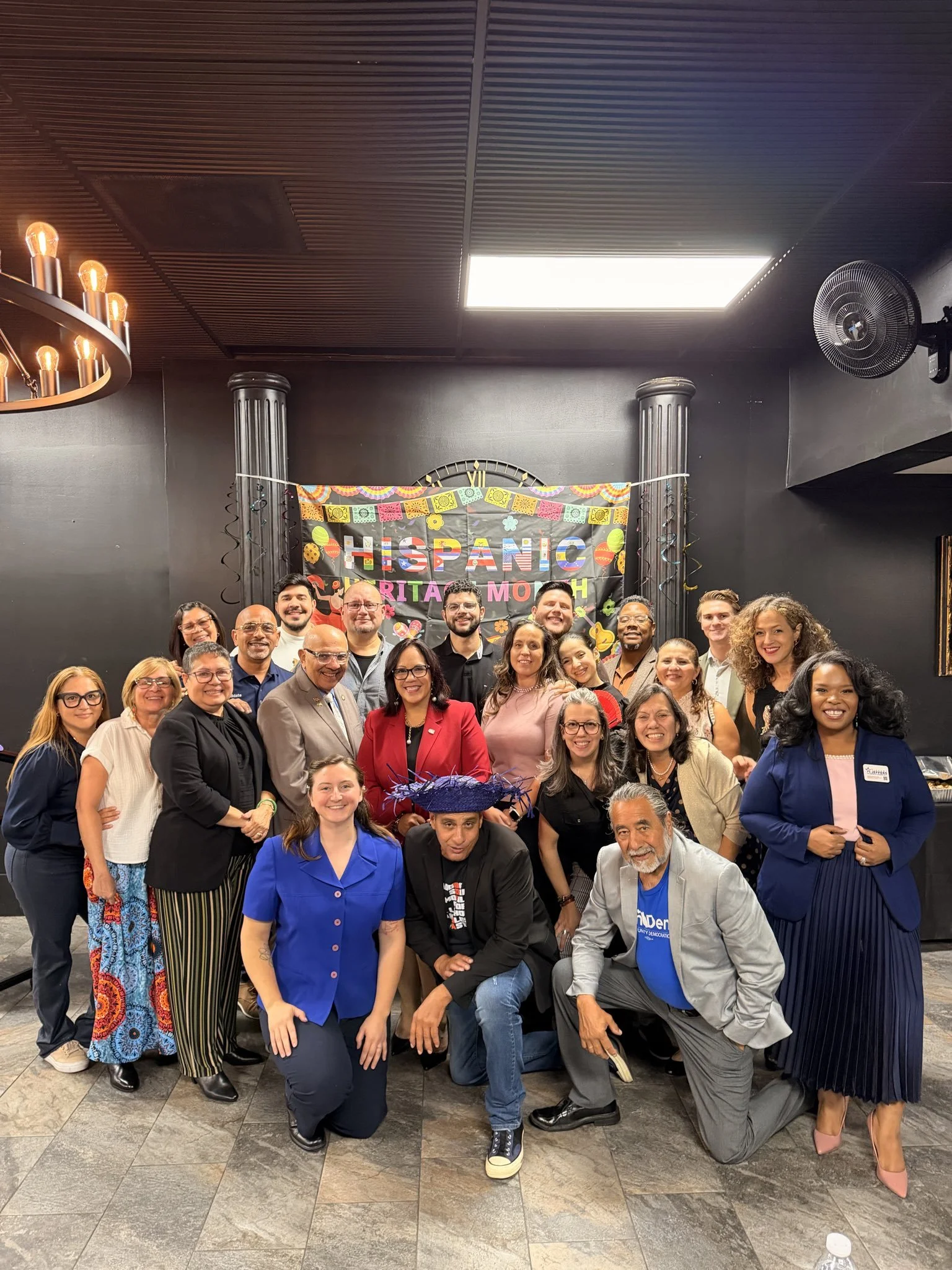 Group of people celebrating a Hispanic event, standing in a room with a decorated backdrop that reads 'HISPANIC' and 'MARITA MO' with colorful decorations.