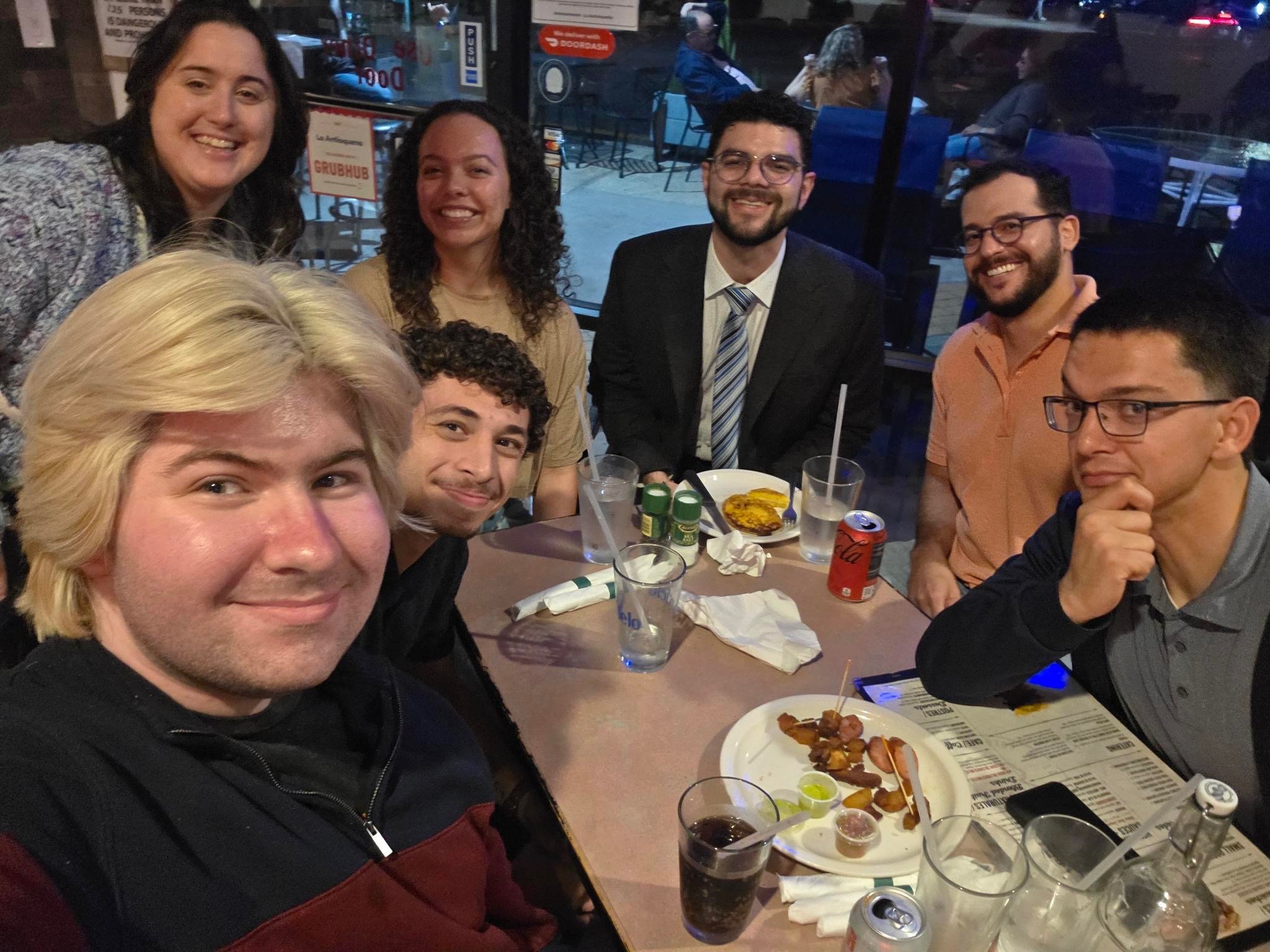 A group of eight people sitting around a restaurant table, smiling for the camera. The table has plates of food, drinks, and condiments. The background shows large windows and some customers outside.