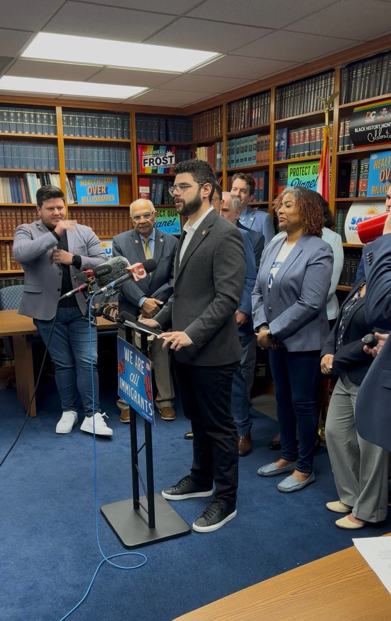 A group of people at a press conference indoors, with bookshelves, signs, and flags in the background, including a man speaking at a podium.